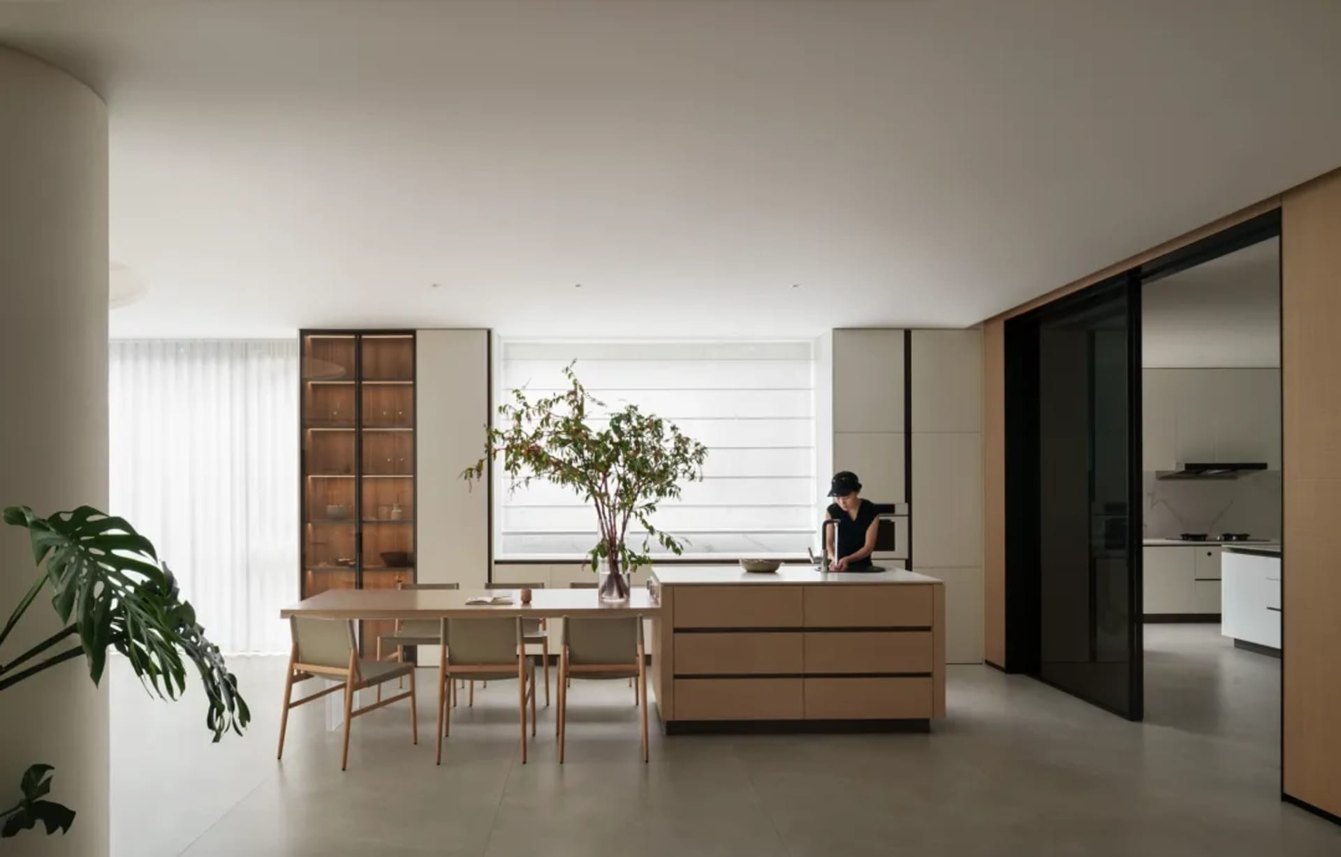 Modern kitchen with wooden cabinets, white walls, and a large window allowing natural light to fill the space, featuring two dining tables and several chairs, with a potted plant adding greenery