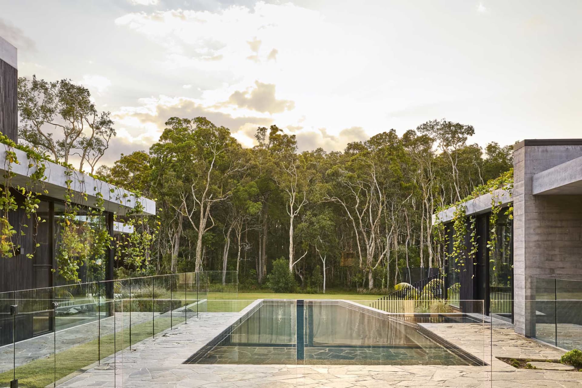 A serene outdoor pool scene with a glass-enclosed pool surrounded by trees and two houses in the background