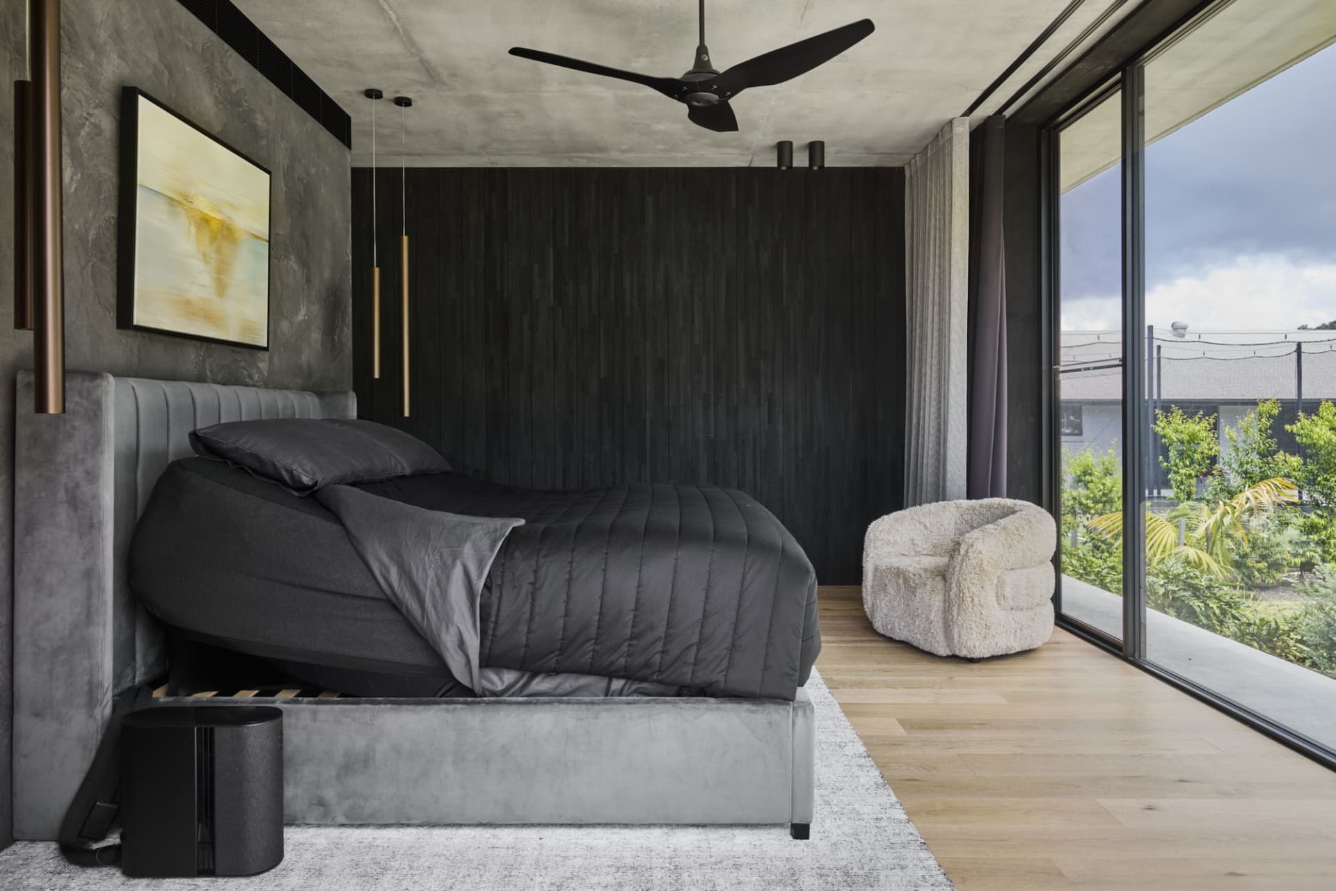 A modern bedroom with a black bed on a wooden floor, gray and white tones, ceiling fans, a chair, and a potted plant