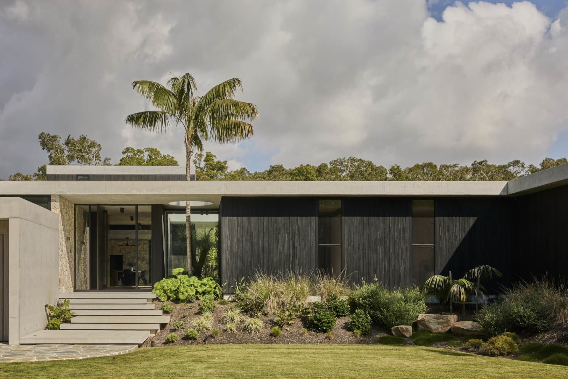 A modern house with a black exterior, featuring a large window and surrounded by palm trees, set in a tropical atmosphere
