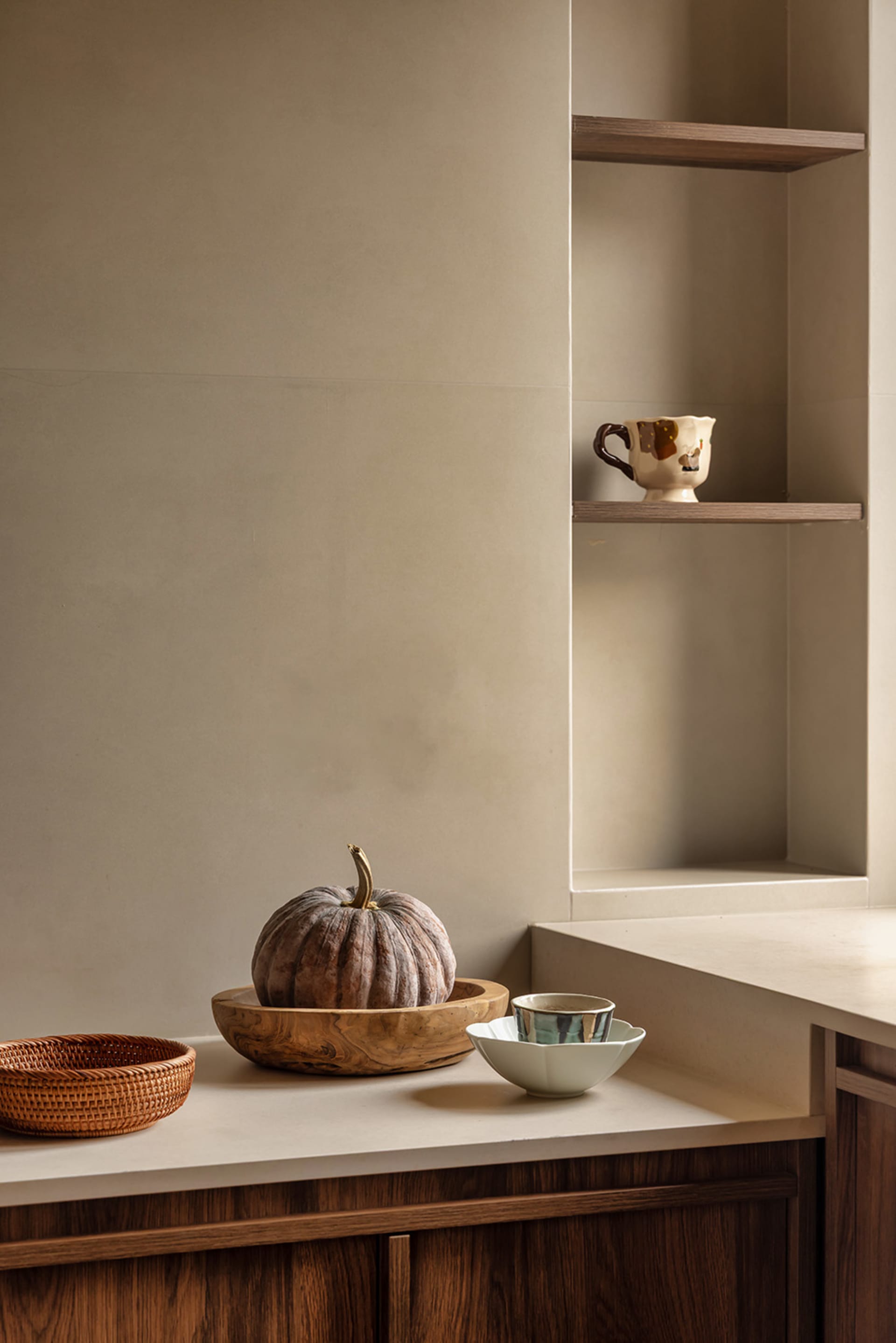 A kitchen countertop with three bowls, two cups, two spoons, and a pumpkin, set against a neutral background