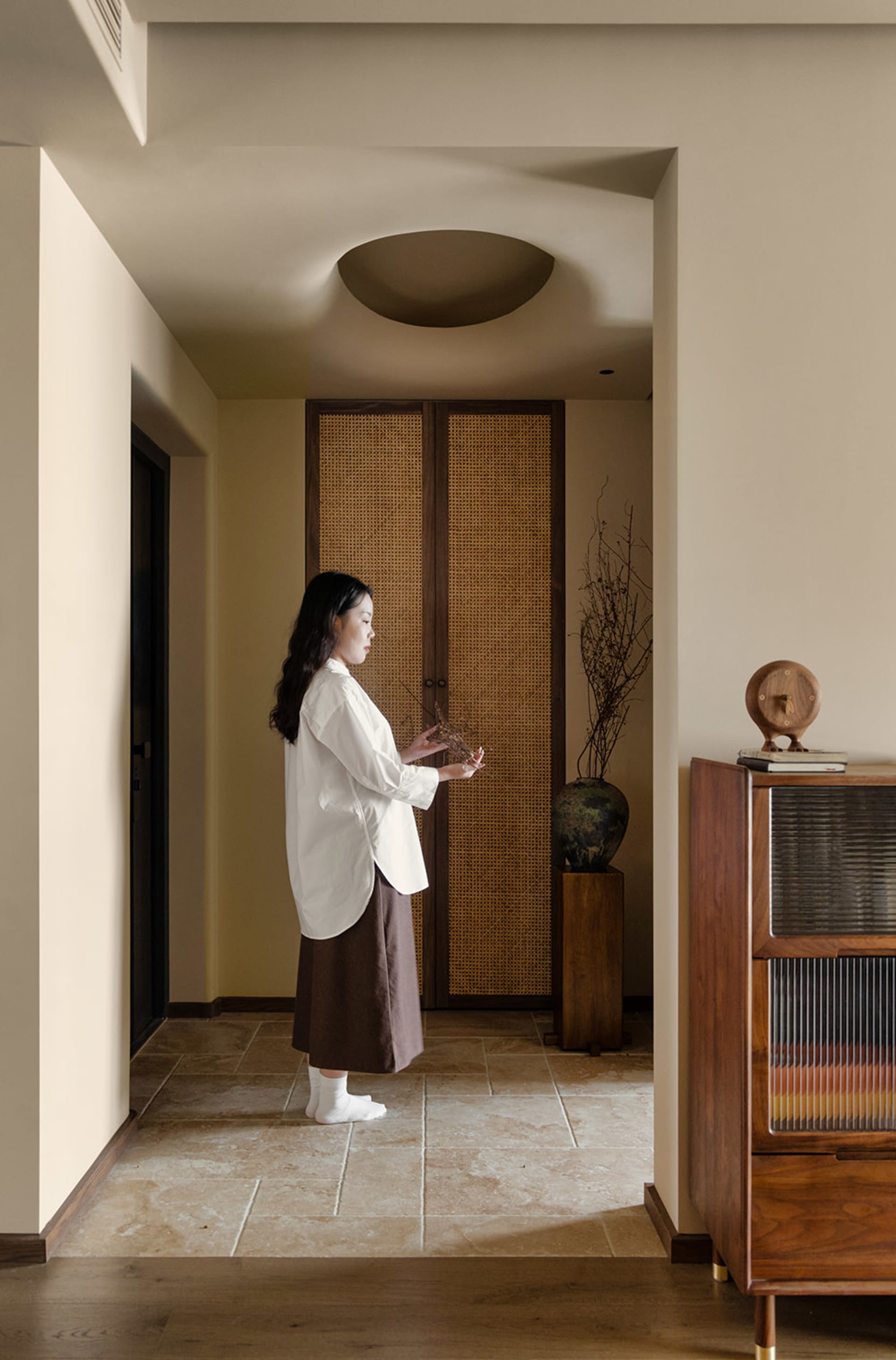 A woman stands in a cozy living room with a wooden table, vase, and books, surrounded by a comfortable atmosphere