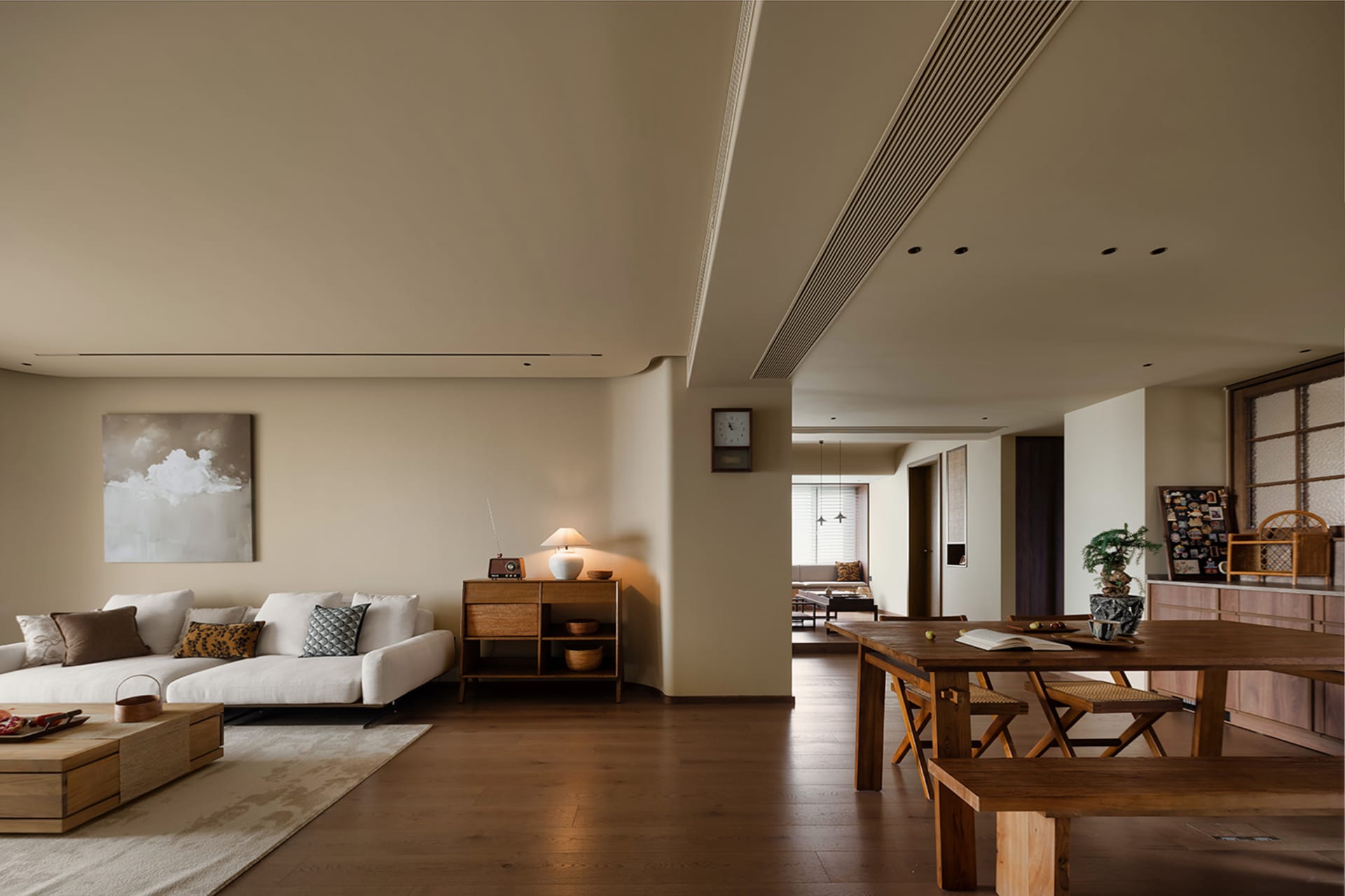 Warm and inviting living space with wooden floors, white walls, and natural light from a large window, featuring two couches, a dining table, and a clock on the wall