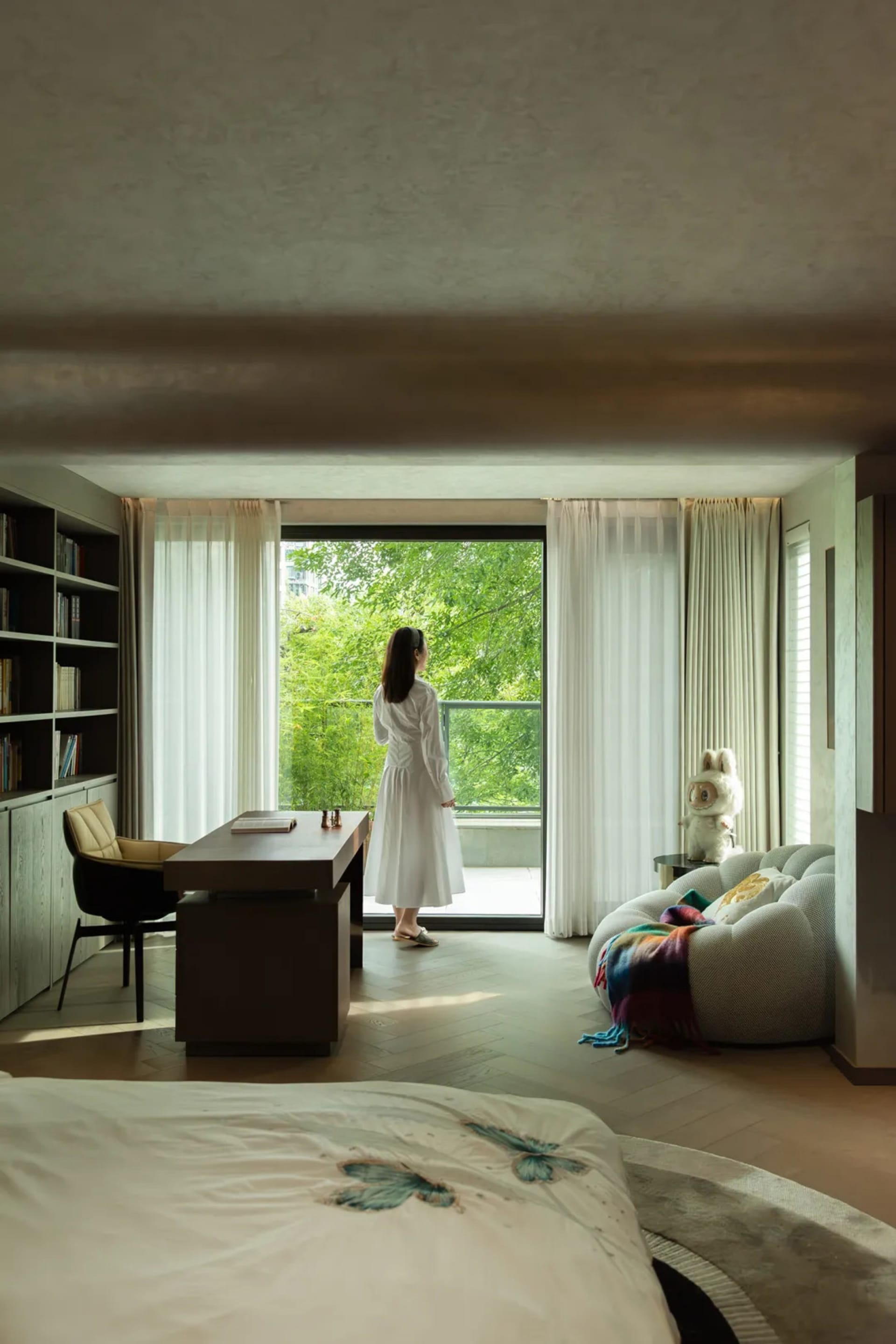 A woman stands near a window in a well-lit room with a wooden floor, two chairs, a couch, and a teddy bear on the floor