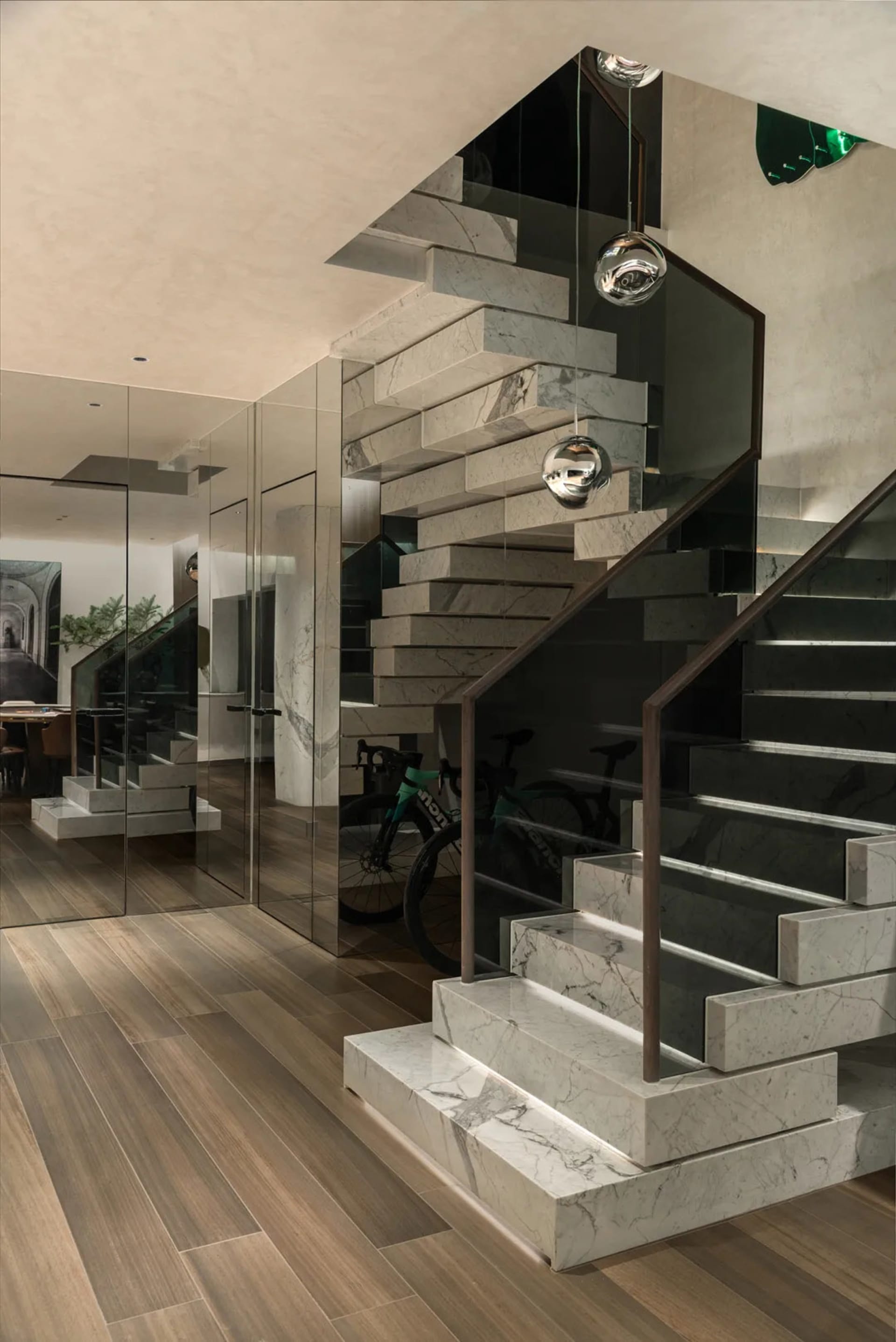 A well-maintained interior staircase with a marble stairwell, glass walls, wooden stairs, and a metal railing, featuring bicycles and a potted plant