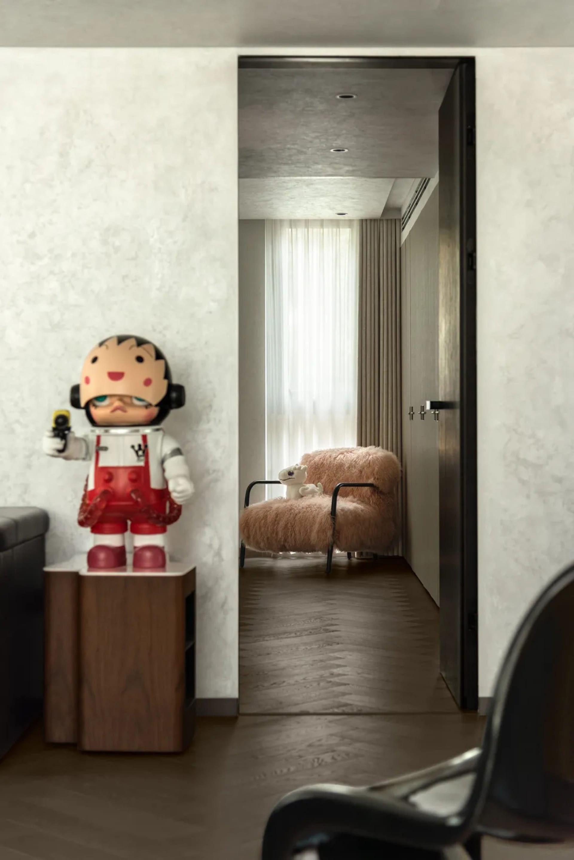 A small living area with a doll on a chair, a teddy bear on the floor, and a couch in the background, creating a cozy and inviting atmosphere