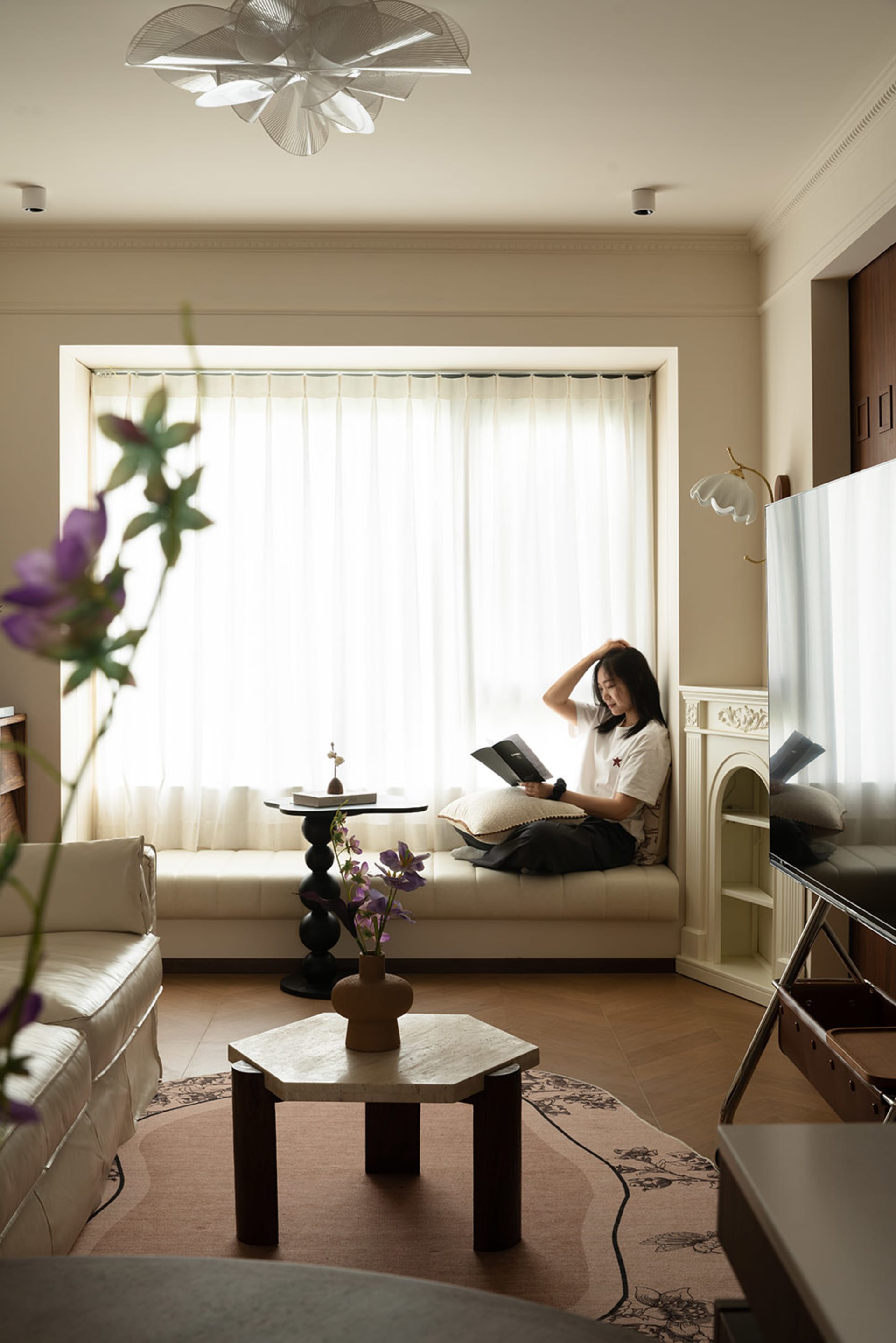 A woman sits on a couch in a cozy living area with a dining table, potted plant, and vases, enjoying a book and the indoor atmosphere