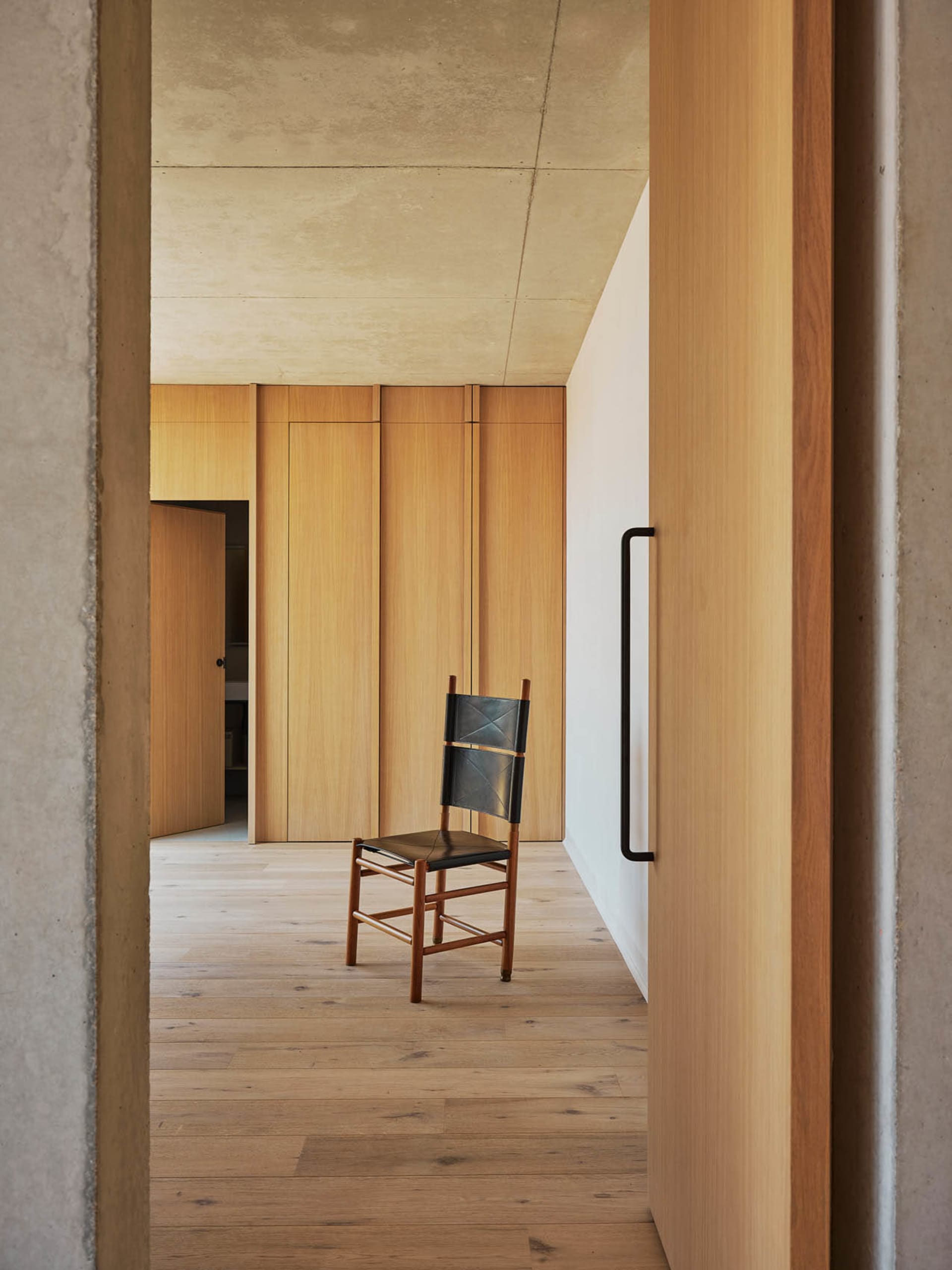 A cozy living area with a wooden floor and a concrete wall, featuring a chair and a backpack, set against a neutral background