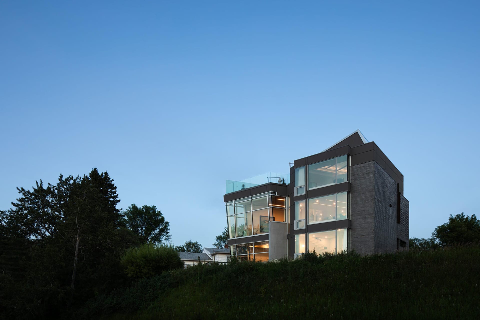 A modern house on a hillside with a combination of glass walls and brick facade, featuring multiple levels, windows, and a rooftop terrace