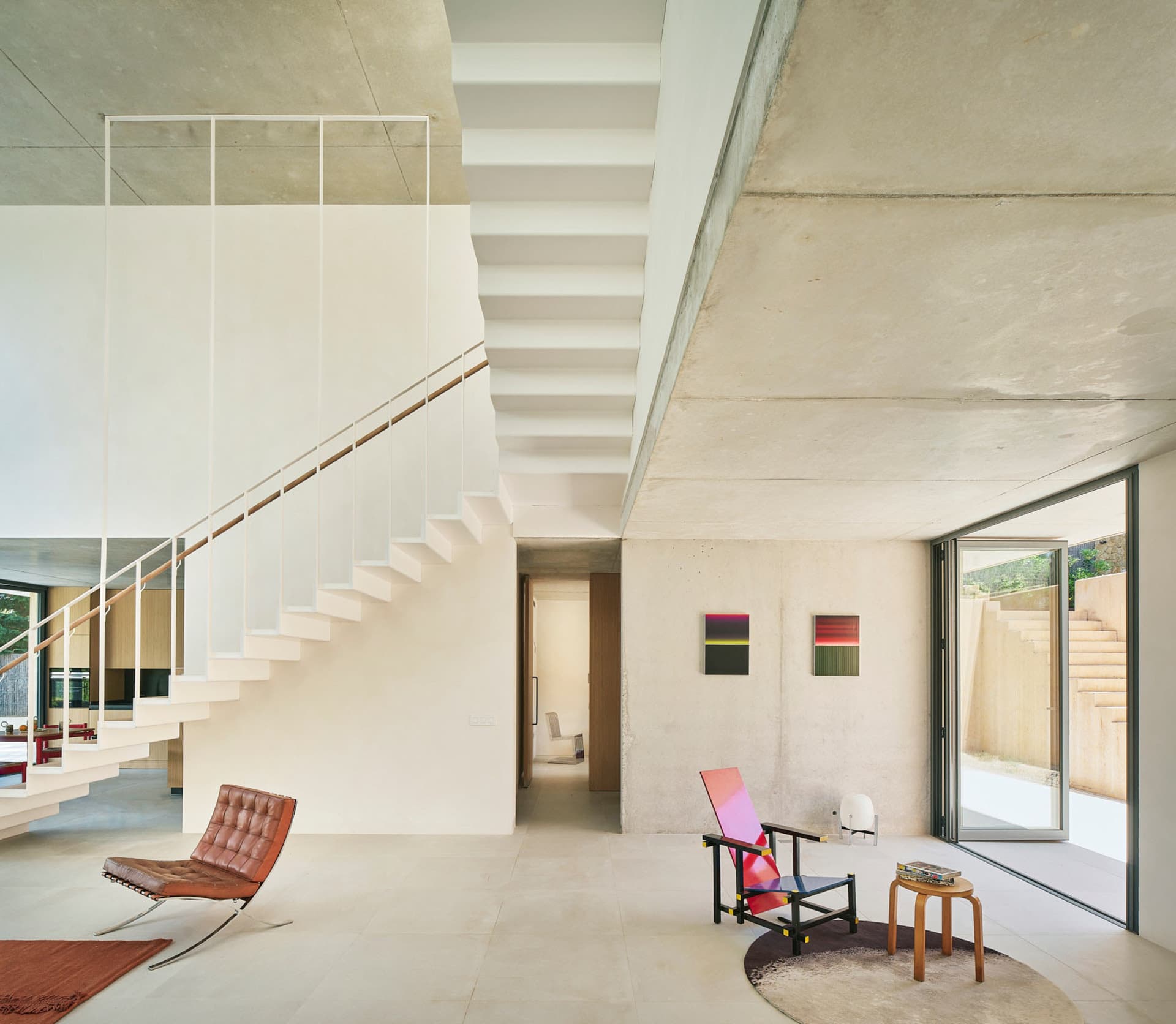 Open modern living area with a white staircase, two chairs, a couch, and natural light from windows, set against a neutral color palette and concrete walls