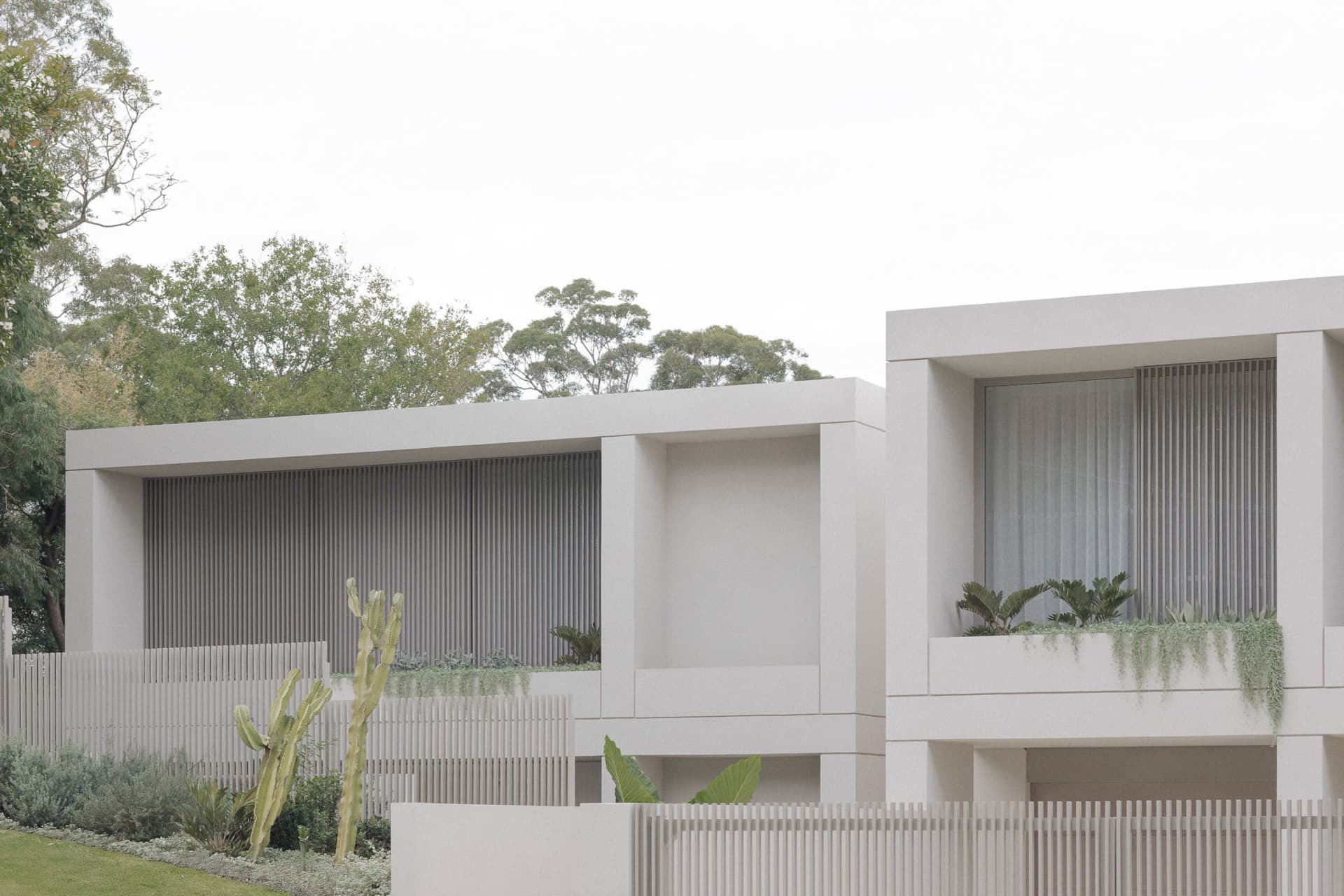 Two distinct houses with a balcony and potted plants, set in a well-maintained residential area with greenery and a sidewalk