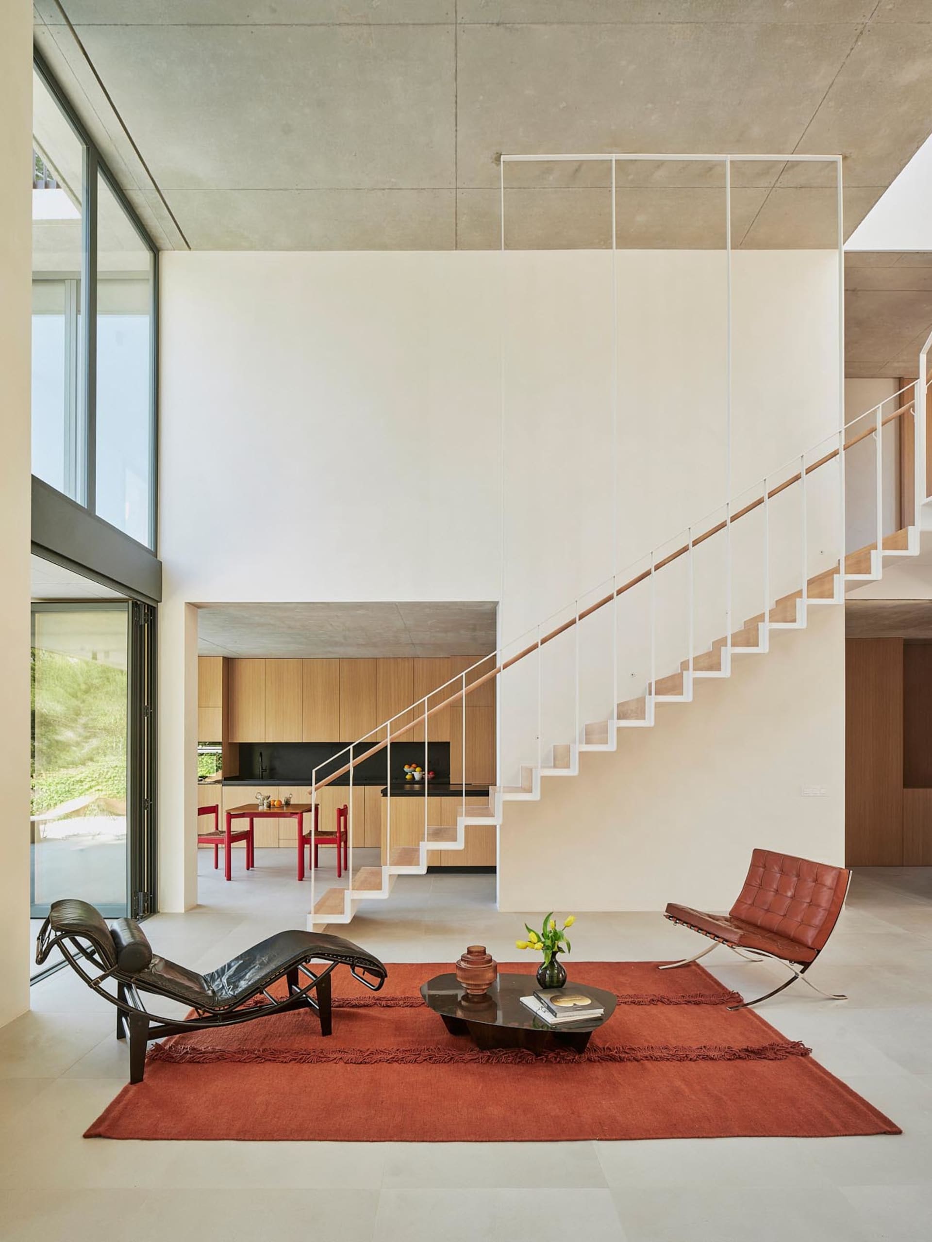 Open modern living area with a white wall backdrop, two staircases, a dining table, a couch, and a potted plant, featuring a clean and minimalist design