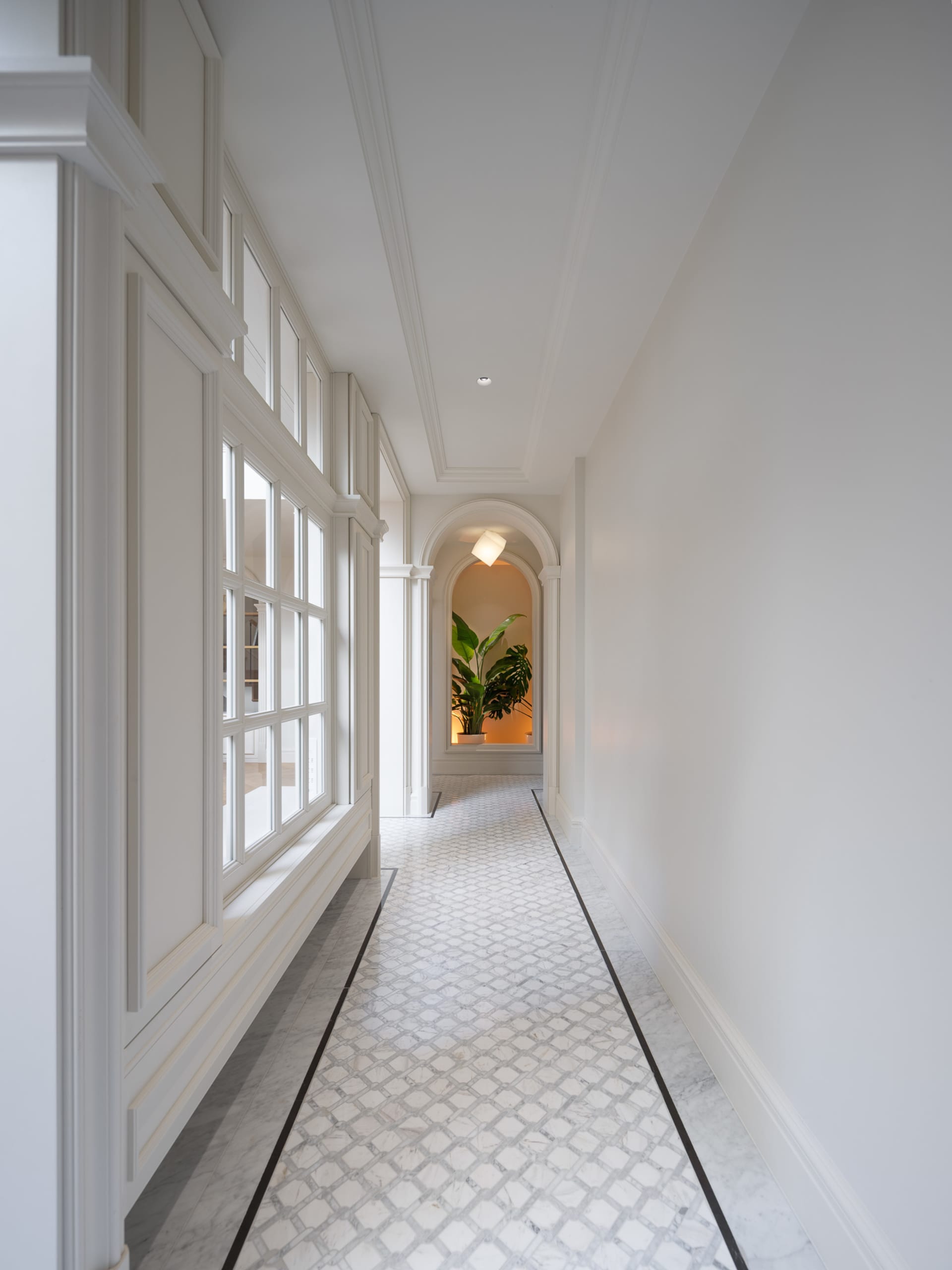 A well-lit hallway with white walls, a tile floor, and two large windows on each side, featuring a potted plant near one of the windows