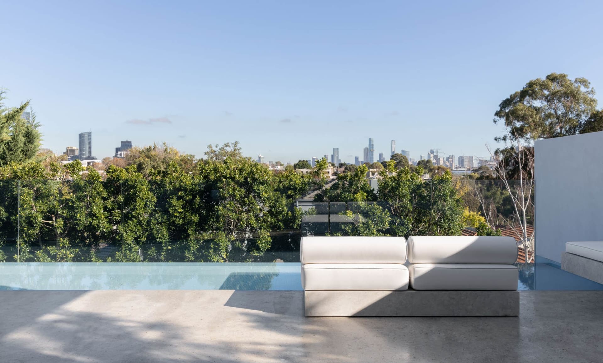 A modern outdoor living area with a white couch on a concrete slab in front of a glass wall with a city view, surrounded by trees