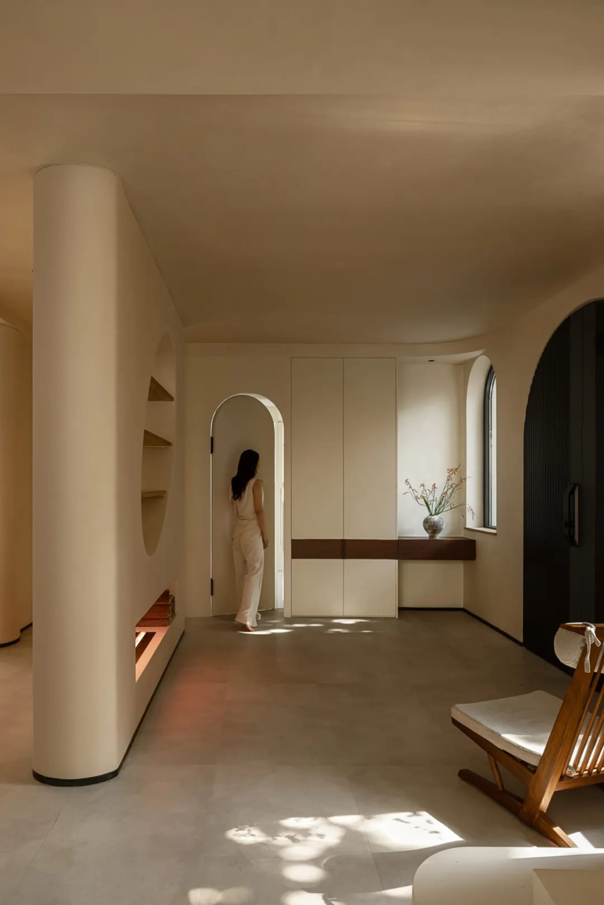 A cozy living area with a woman standing in a hallway between two rooms, featuring two chairs and a potted plant, creating a warm and inviting atmosphere