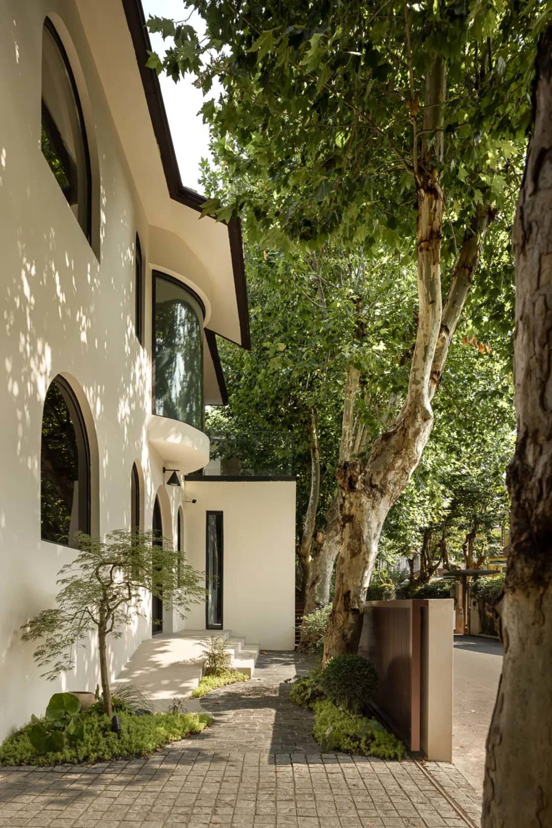 A modern house with a white exterior, surrounded by trees, featuring large windows, potted plants, and a bench, conveying a sense of comfort and relaxation