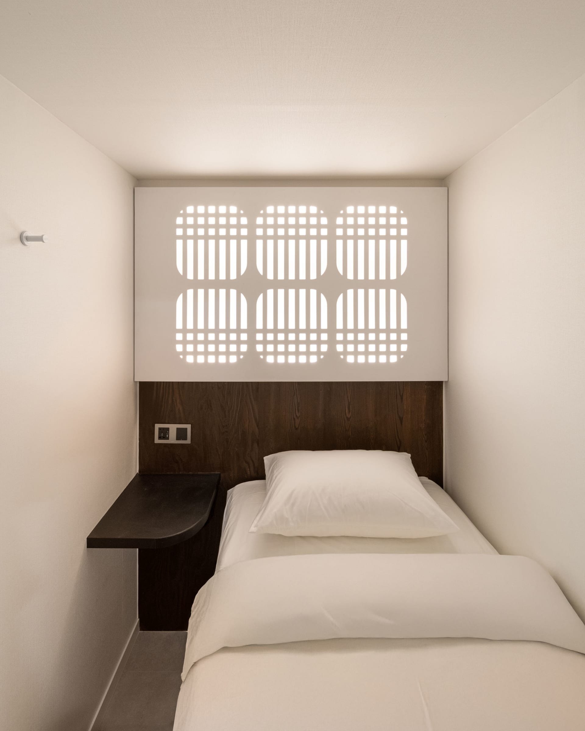 A modern bedroom with a white comforter and brown headboard, a desk, and a chair, well-lit by natural light through a window