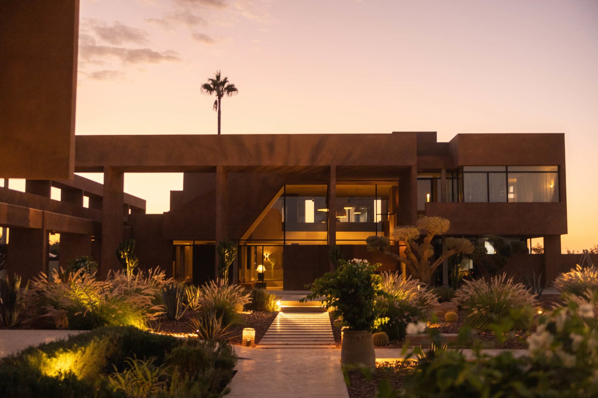 A modern desert-style house with a mix of stone and wood materials, surrounded by palm trees and well-lit with natural light from the windows