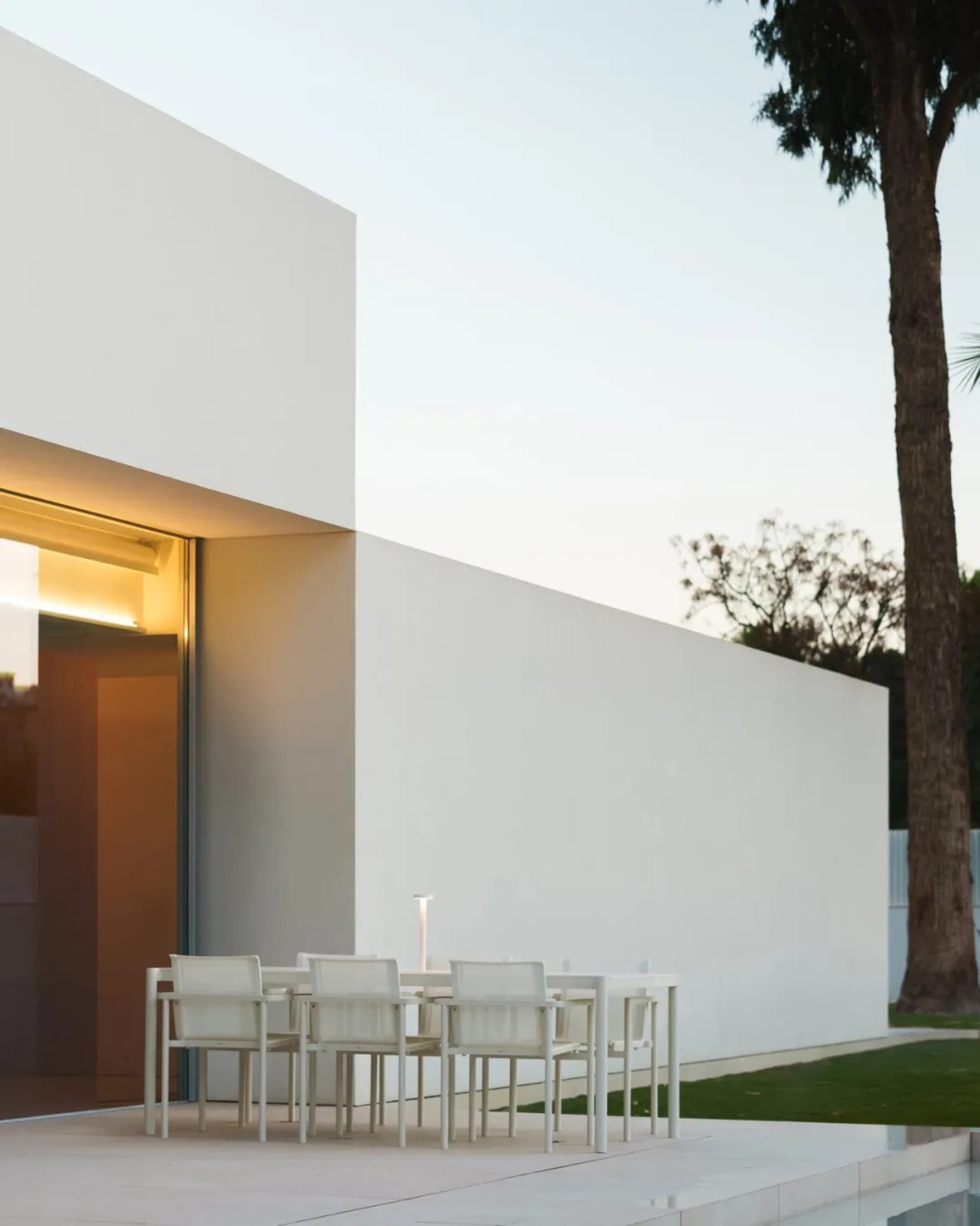 A modern outdoor patio with a dining table and white chairs, surrounded by a white building and a potted plant, creating a comfortable and inviting atmosphere