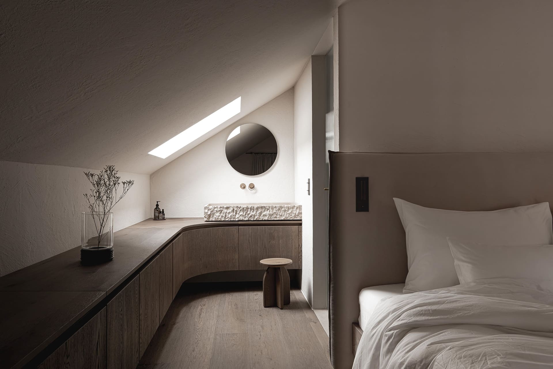 Dimly lit bedroom with a bed, sink, and mirror, featuring wood materials and neutral colors