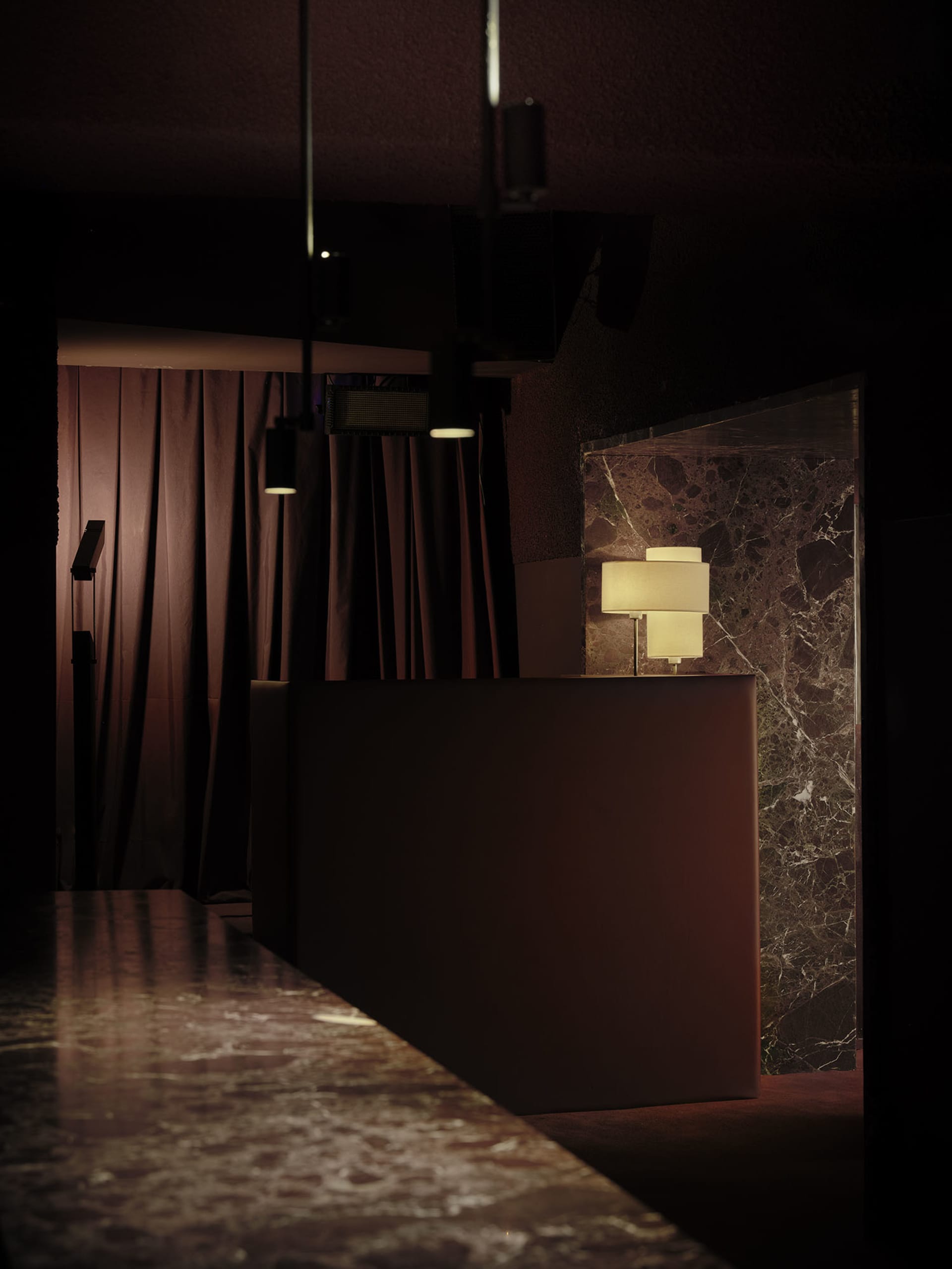 Dimly lit hotel lobby with a marble countertop, a brown table, and a lamp, set against a neutral background