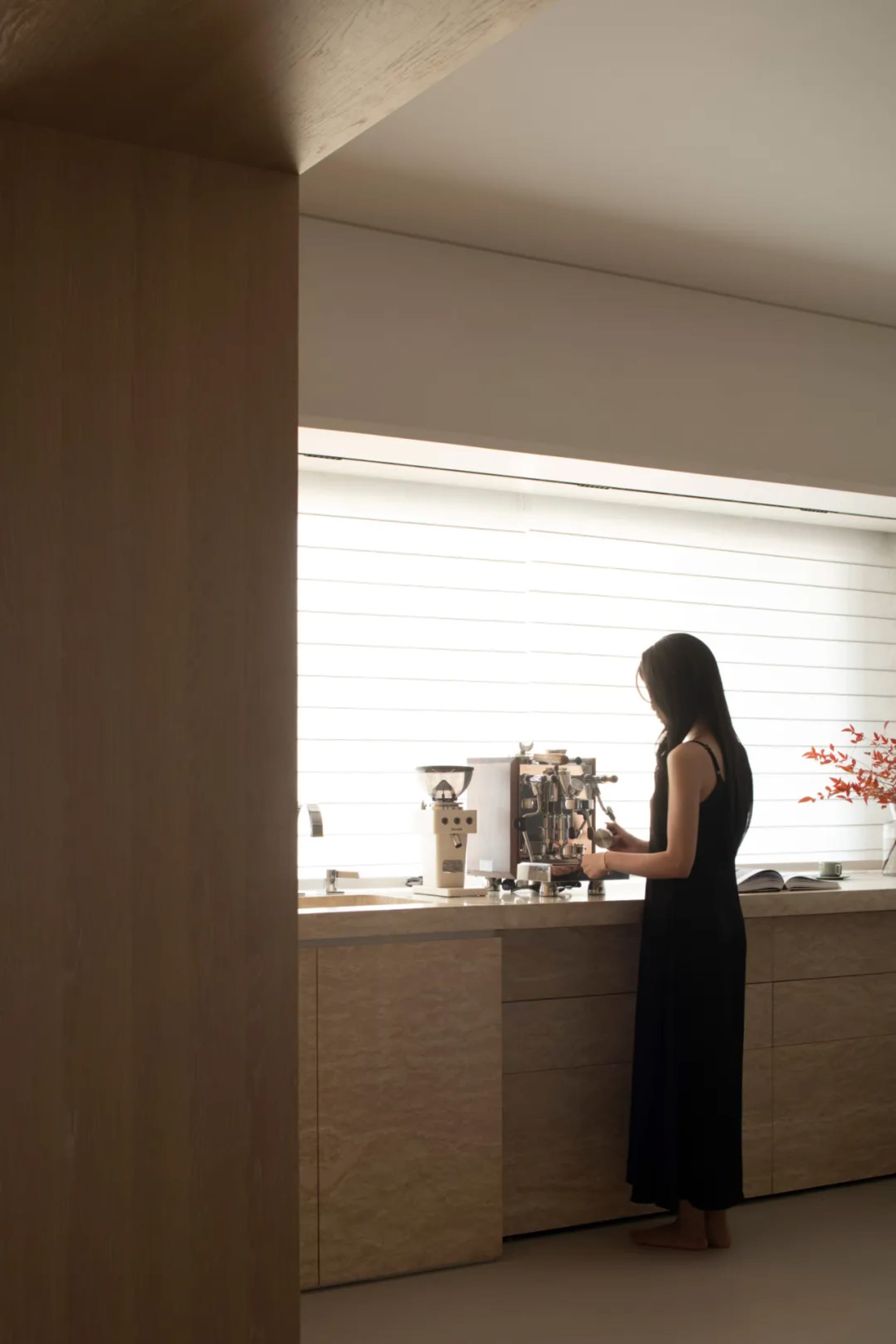 A woman stands in a home kitchen, preparing a drink near a sink, refrigerator, and potted plant, with a casual and homey atmosphere