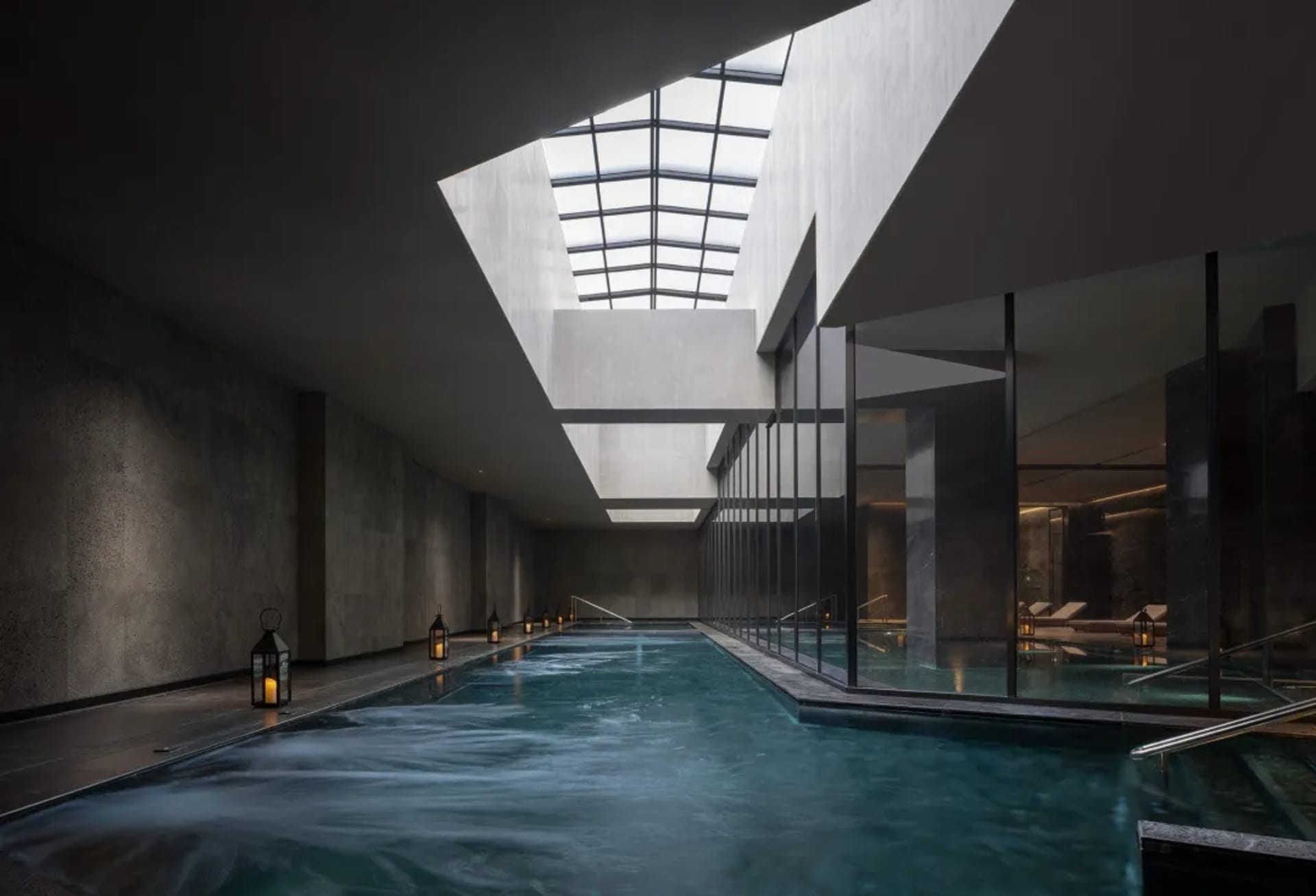 A serene and inviting pool area with a glass ceiling, surrounded by a stone building and well-lit with several windows