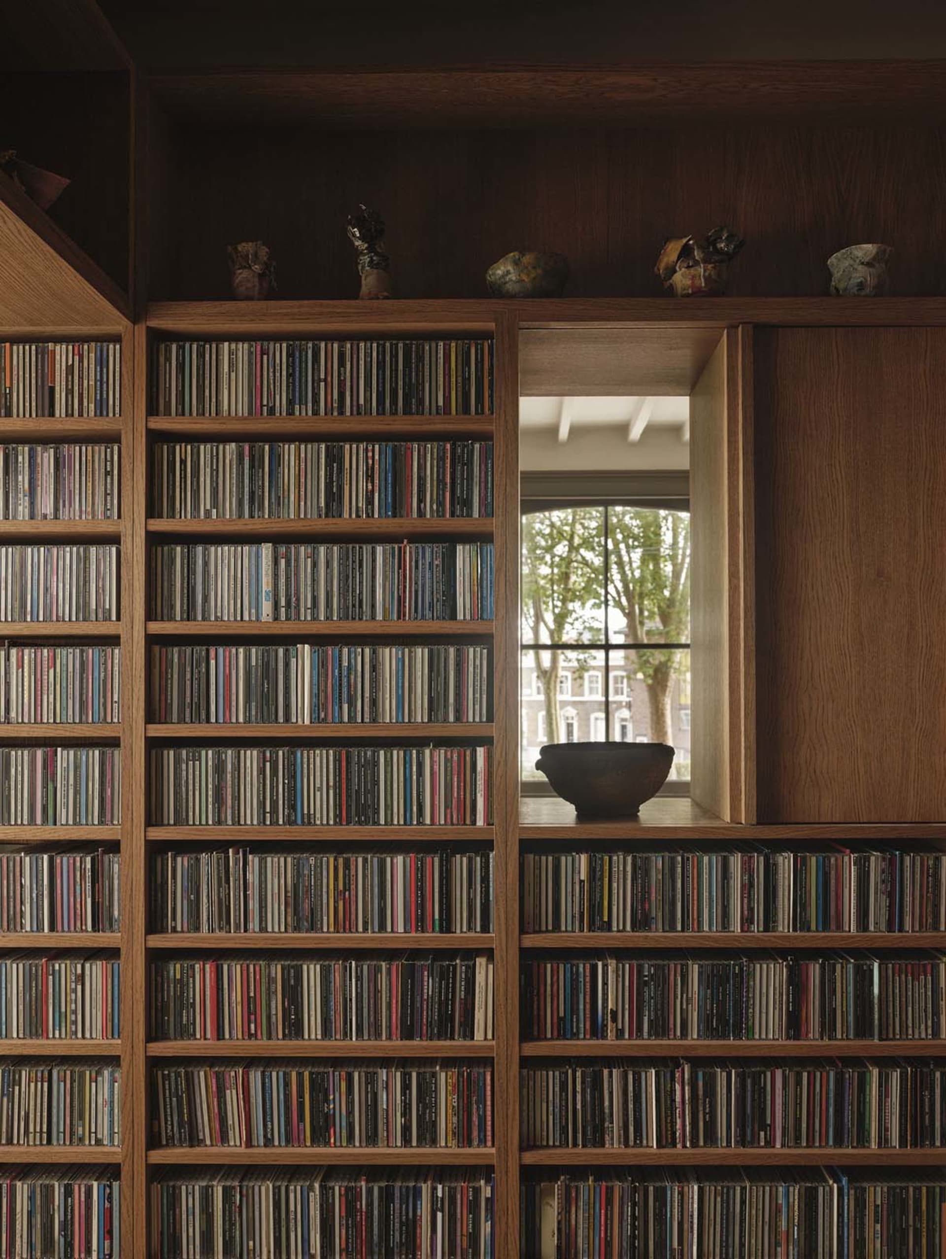 A cozy room with a wooden bookshelf near a window, holding various CDs and a bowl, creating a warm and inviting atmosphere
