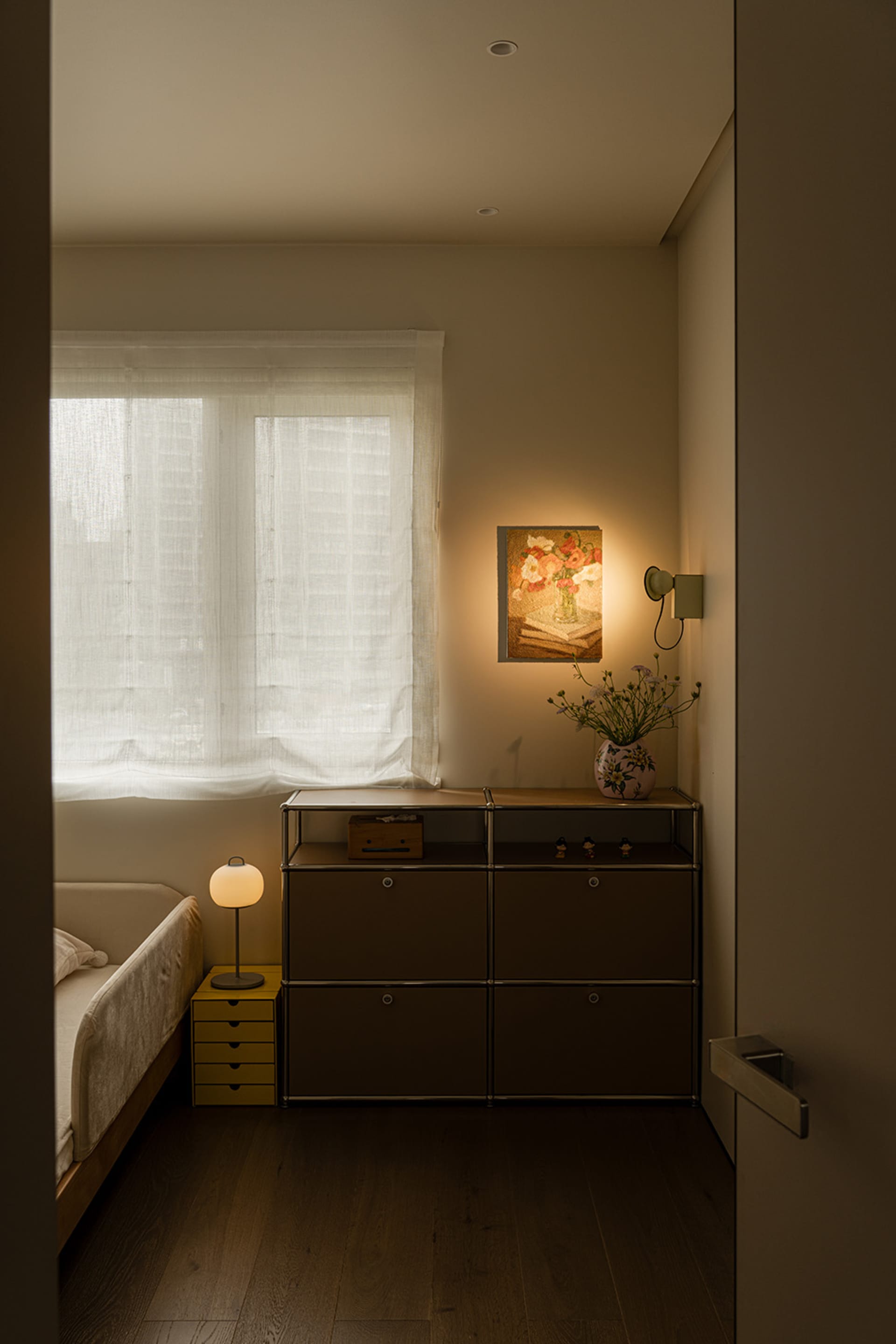 Cozy bedroom with a bed against the wall, a dresser next to it, and a lamp on the floor, in a dimly lit space