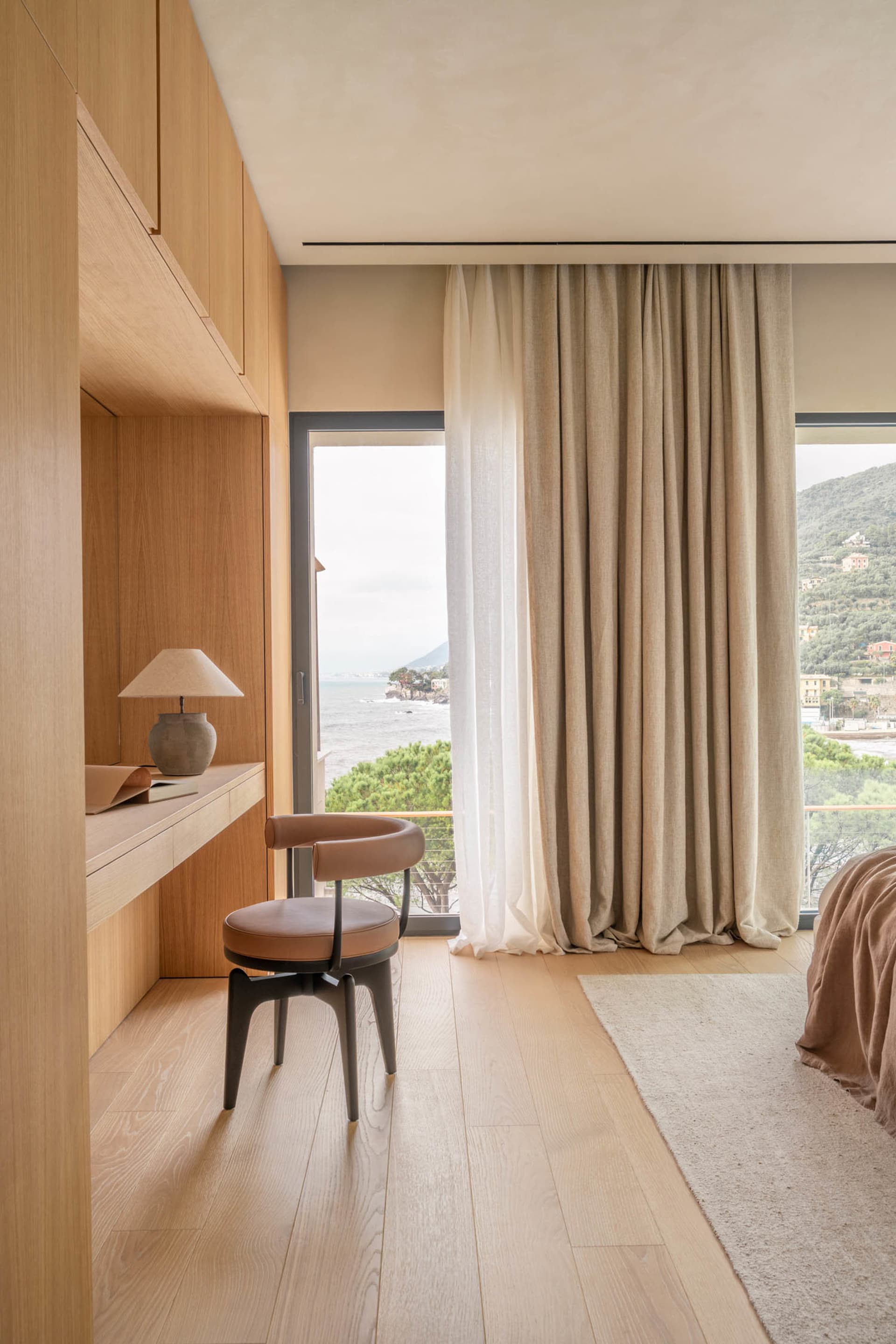 A modern bedroom with a bed, chair, and desk, featuring a large window with natural light and a view of the outdoors