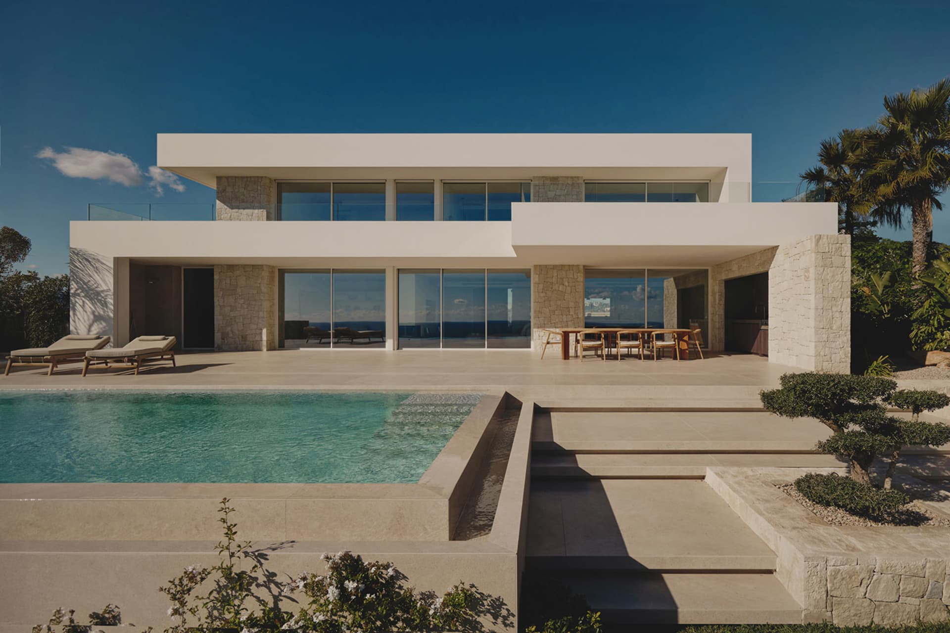 A modern house with a large glass wall, pool, and patio area, surrounded by white surfaces and natural light