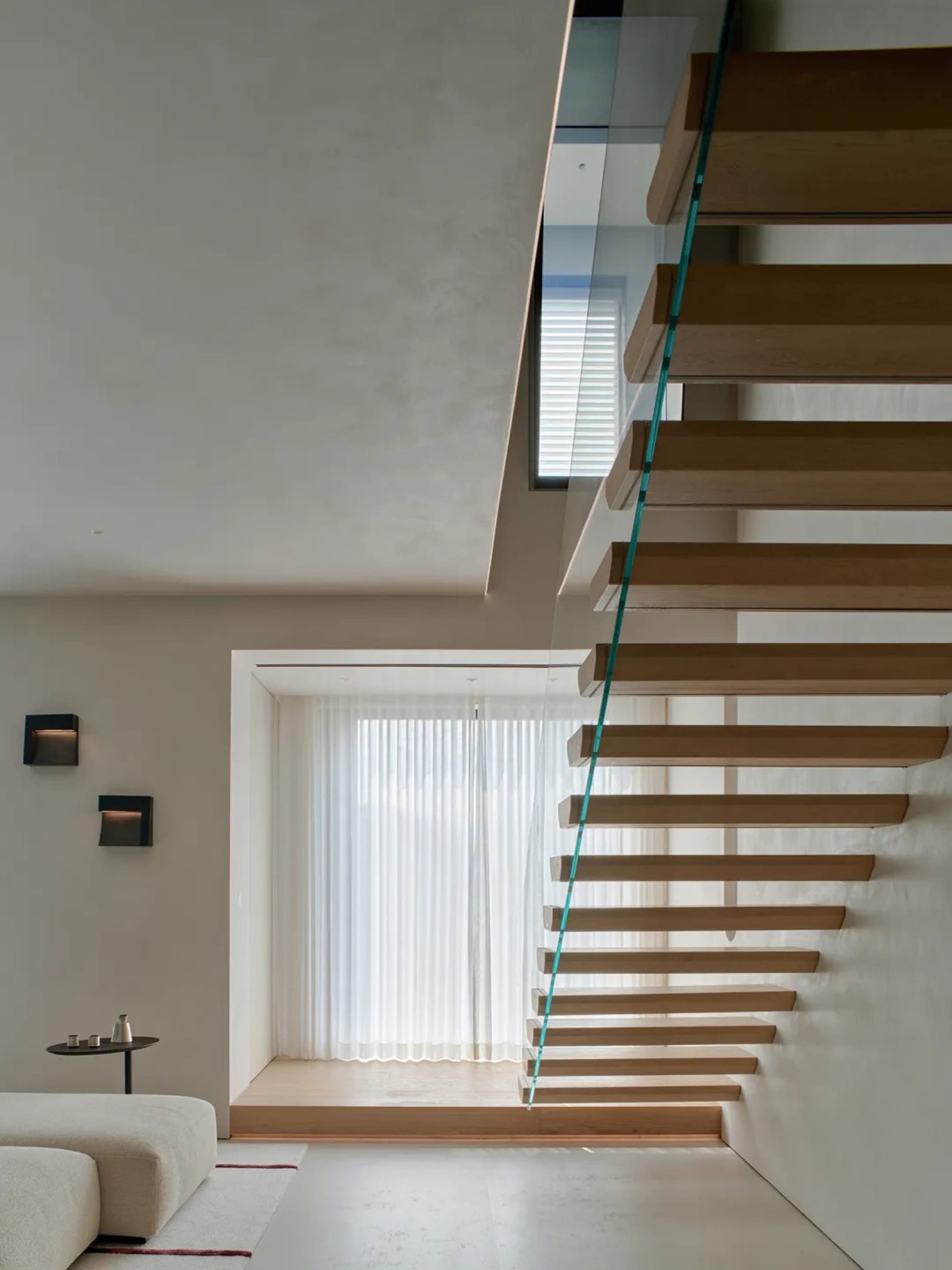 A wooden staircase with a glass railing in a room with a window, lit by natural light