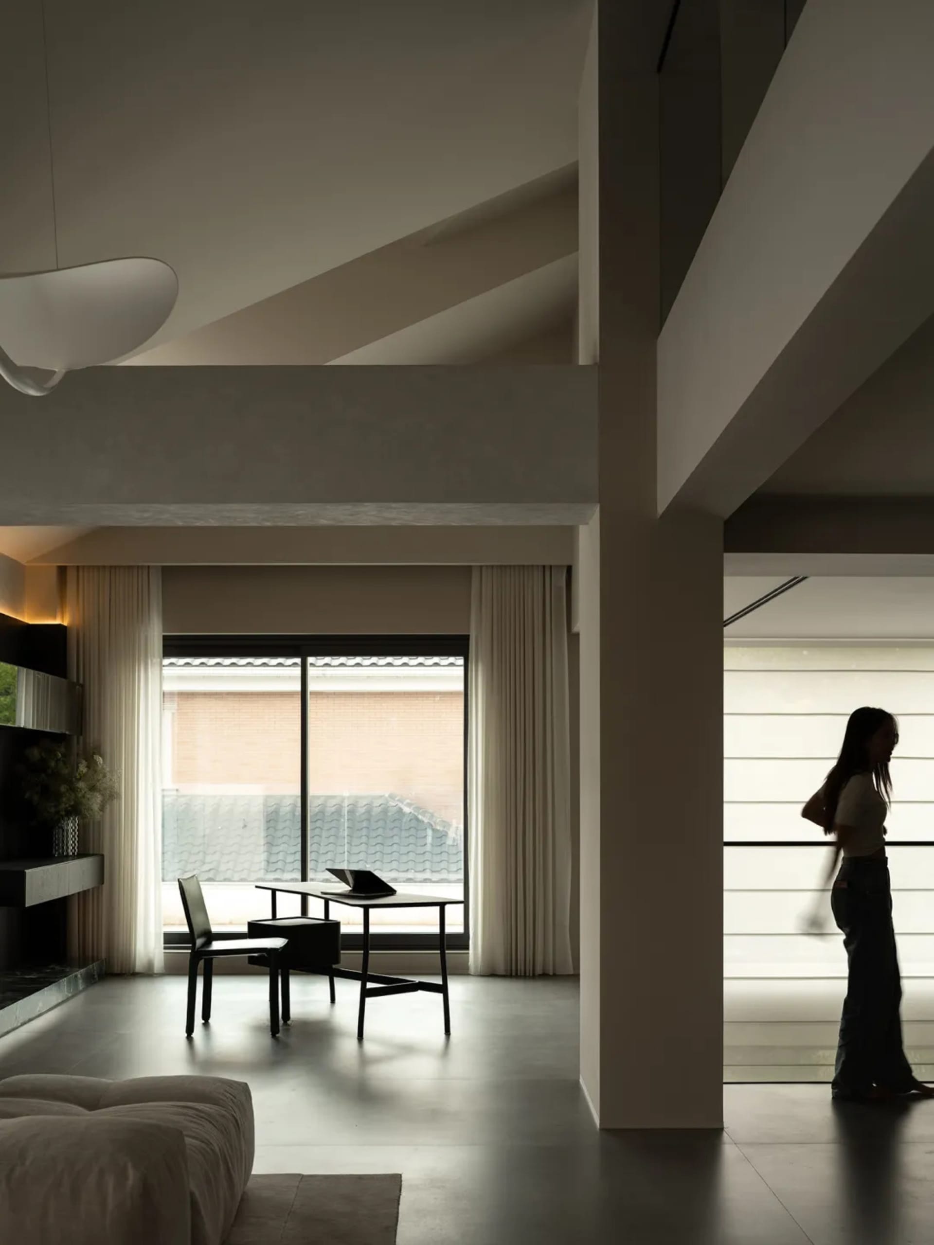 Woman walking towards a dining table in a modern living room with a couch, lamp, and white and grey colors, dim lighting creating a cozy atmosphere