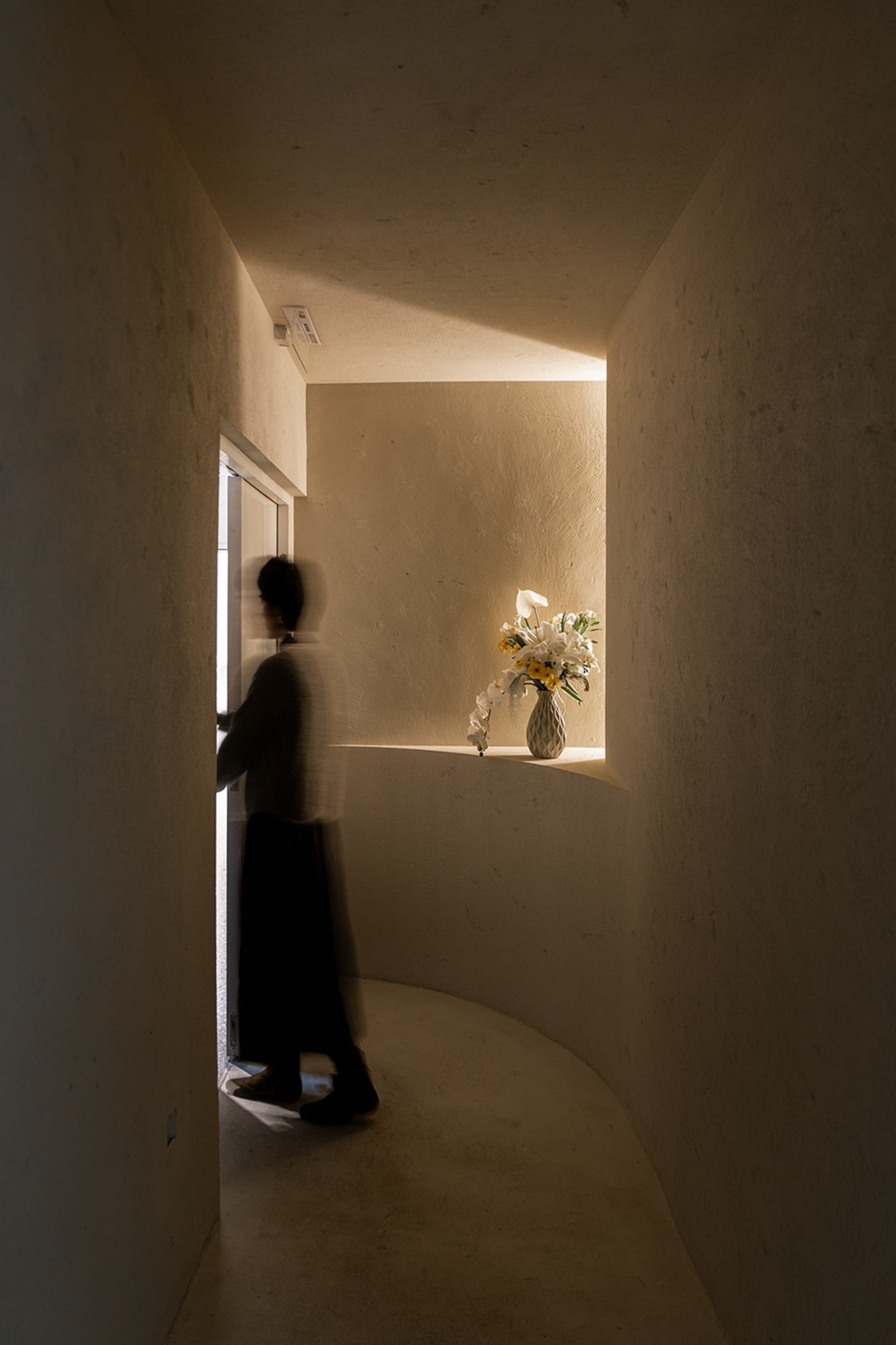 A calm hallway with a woman walking down it, featuring concrete walls, a vase with flowers on a shelf, and a serene atmosphere