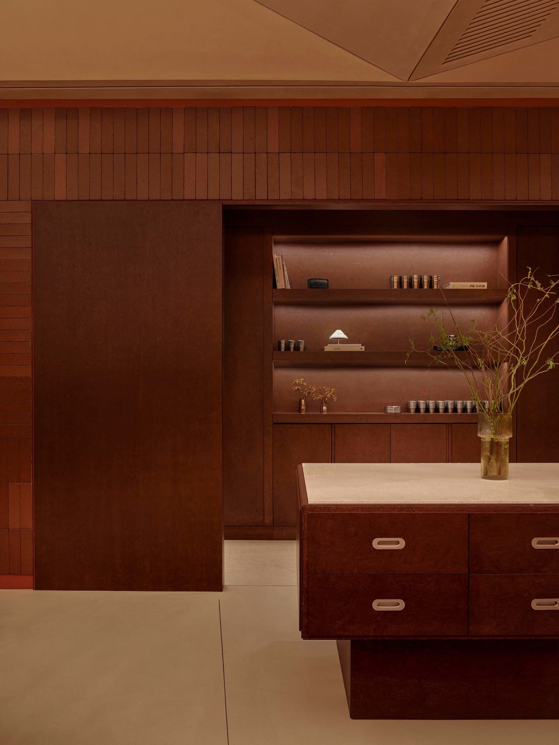Bathroom with a wooden cabinet, marble counter, and a vase of flowers, set against a brown color scheme with a potted plant and dim lighting