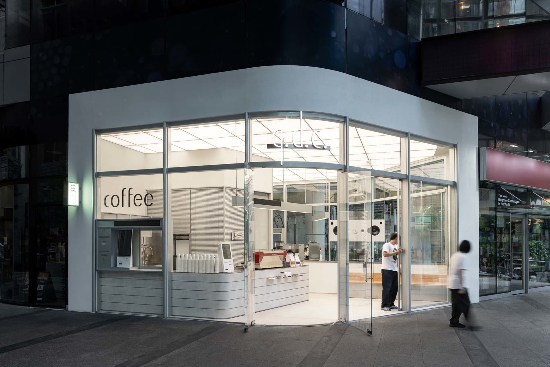 A modern coffee shop with a glass front, white interior, and natural light, featuring appliances, furniture, and a welcoming atmosphere