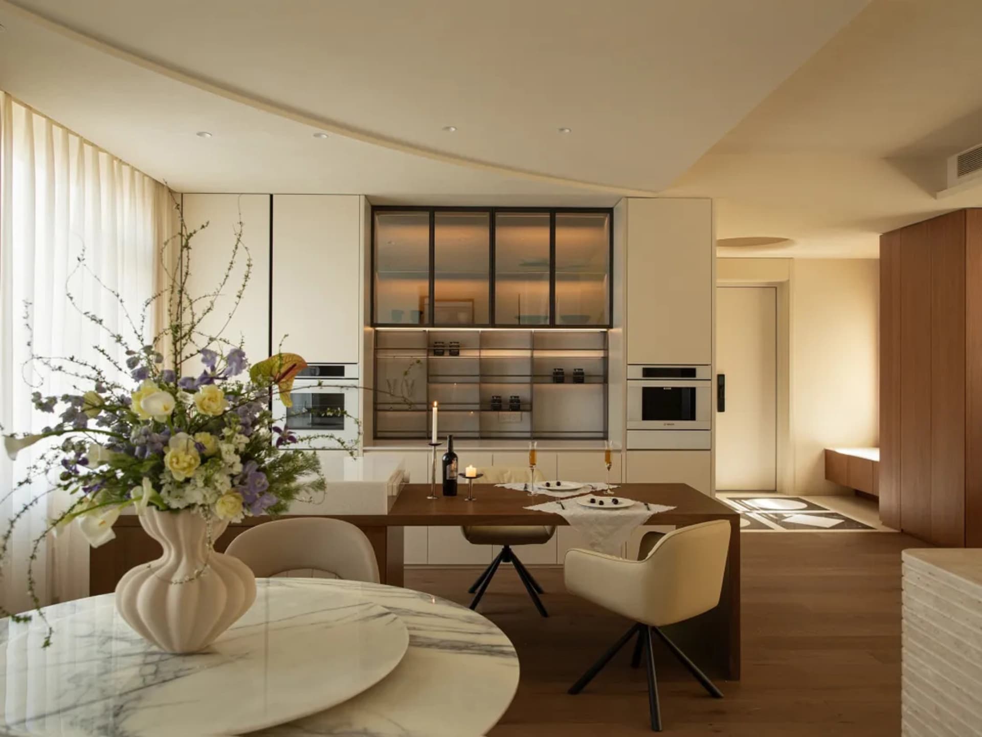 Modern kitchen with a wooden dining table, chairs, and a vase, surrounded by a refrigerator, oven, and microwave, with soft and warm lighting