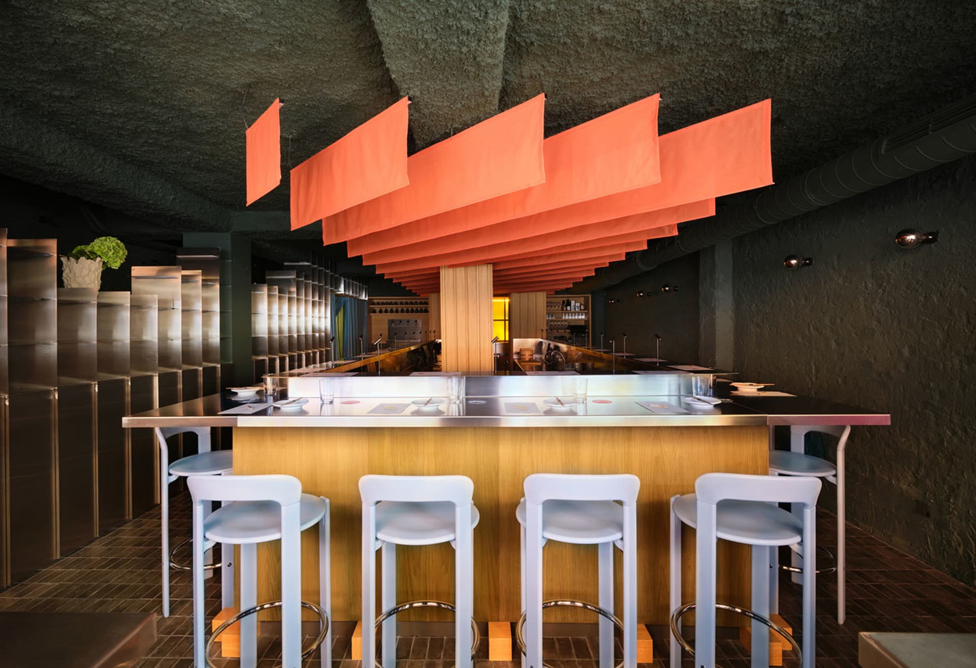 Cozy restaurant with a wooden bar, metal counter, and white stools, featuring a mix of natural and artificial lighting and comfortable seating for guests