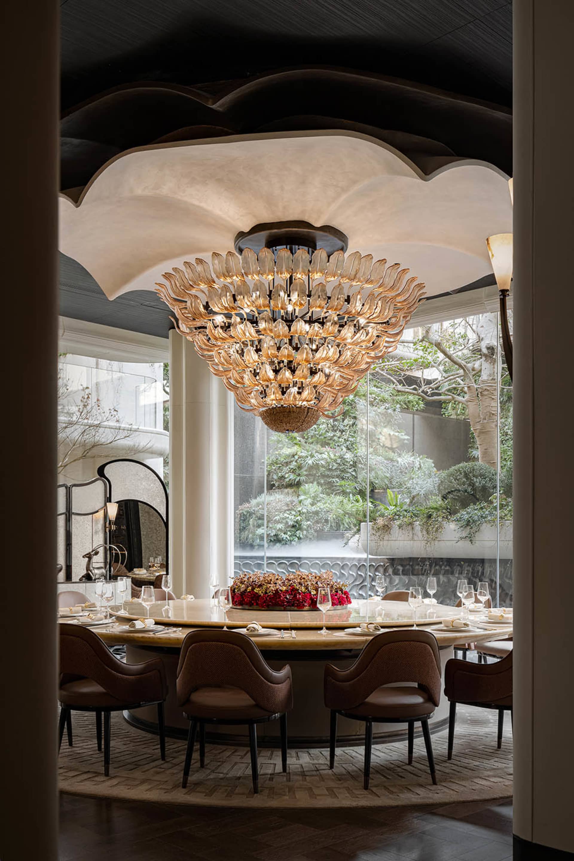 Elegant dining room with a semi-circular seating arrangement, a large chandelier, and potted plants, set for a meal with wine glasses, forks, knives, and spoons