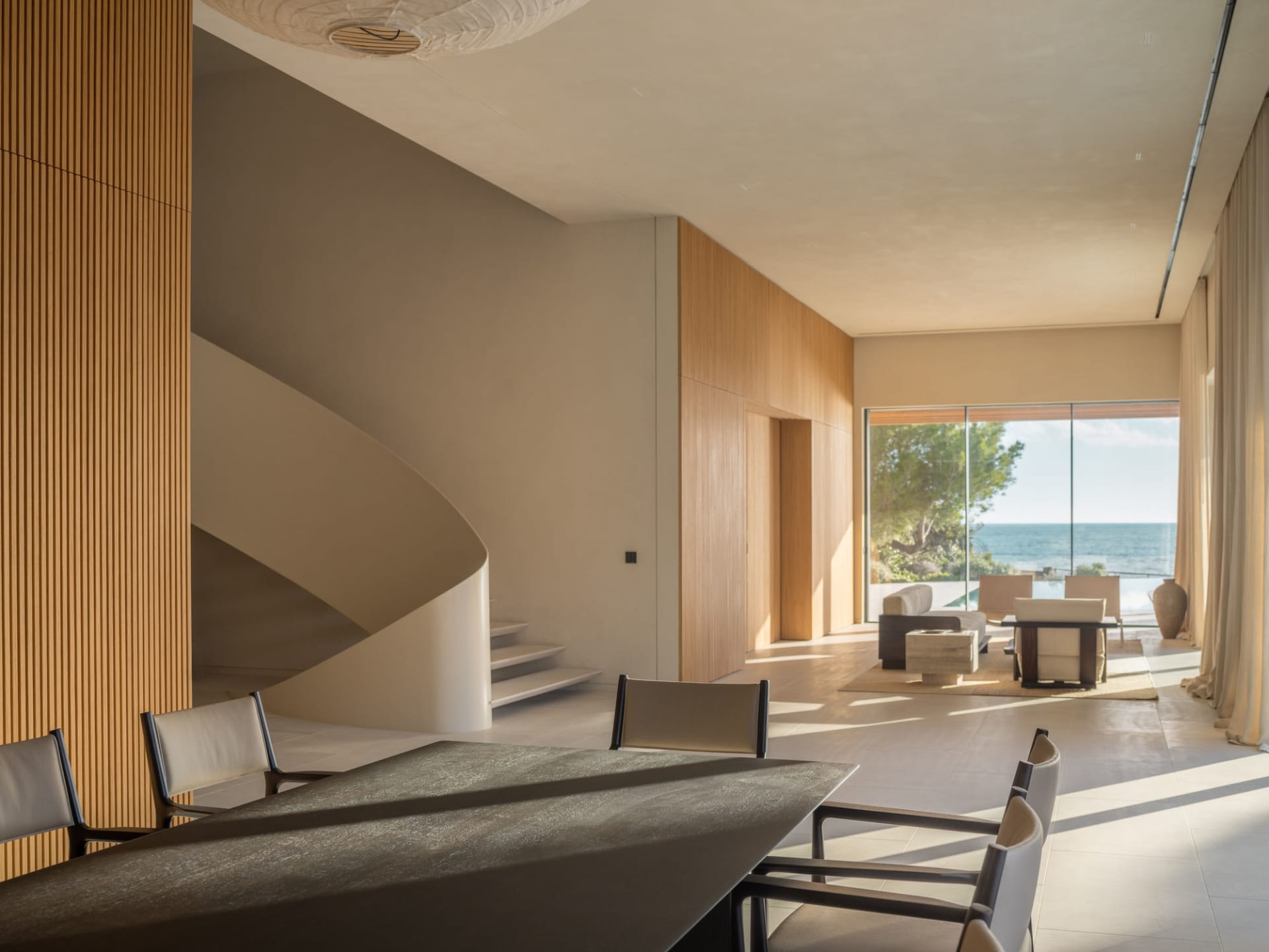 Cozy living room with a dining table, chairs, and a couch arranged around the table, a spiral staircase, and a large window with natural light