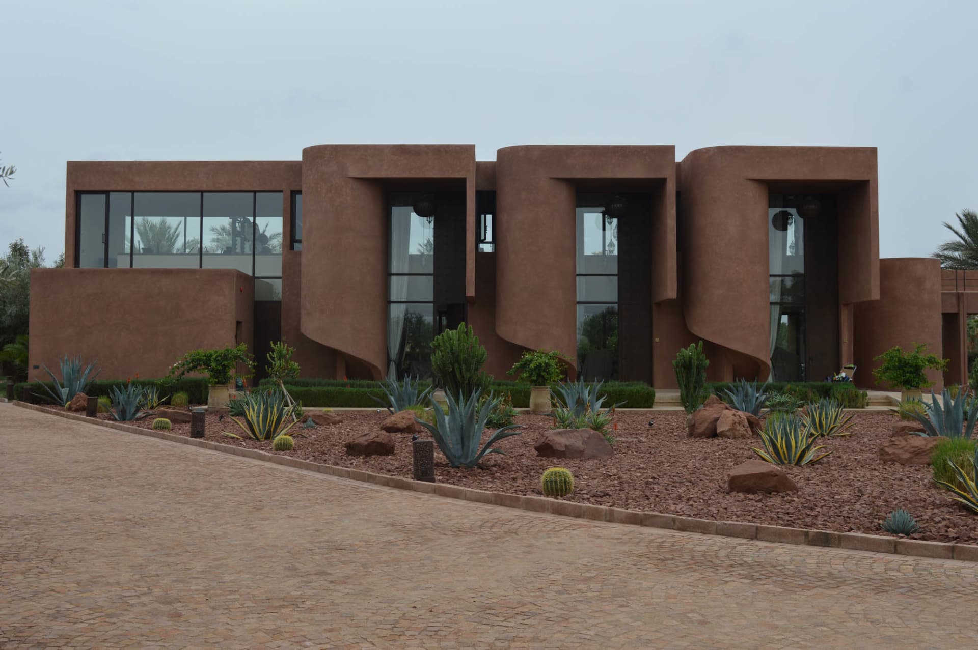 A modern building with a unique design, surrounded by a lush courtyard with cacti and palm trees, featuring red and brown colors and a curved structure