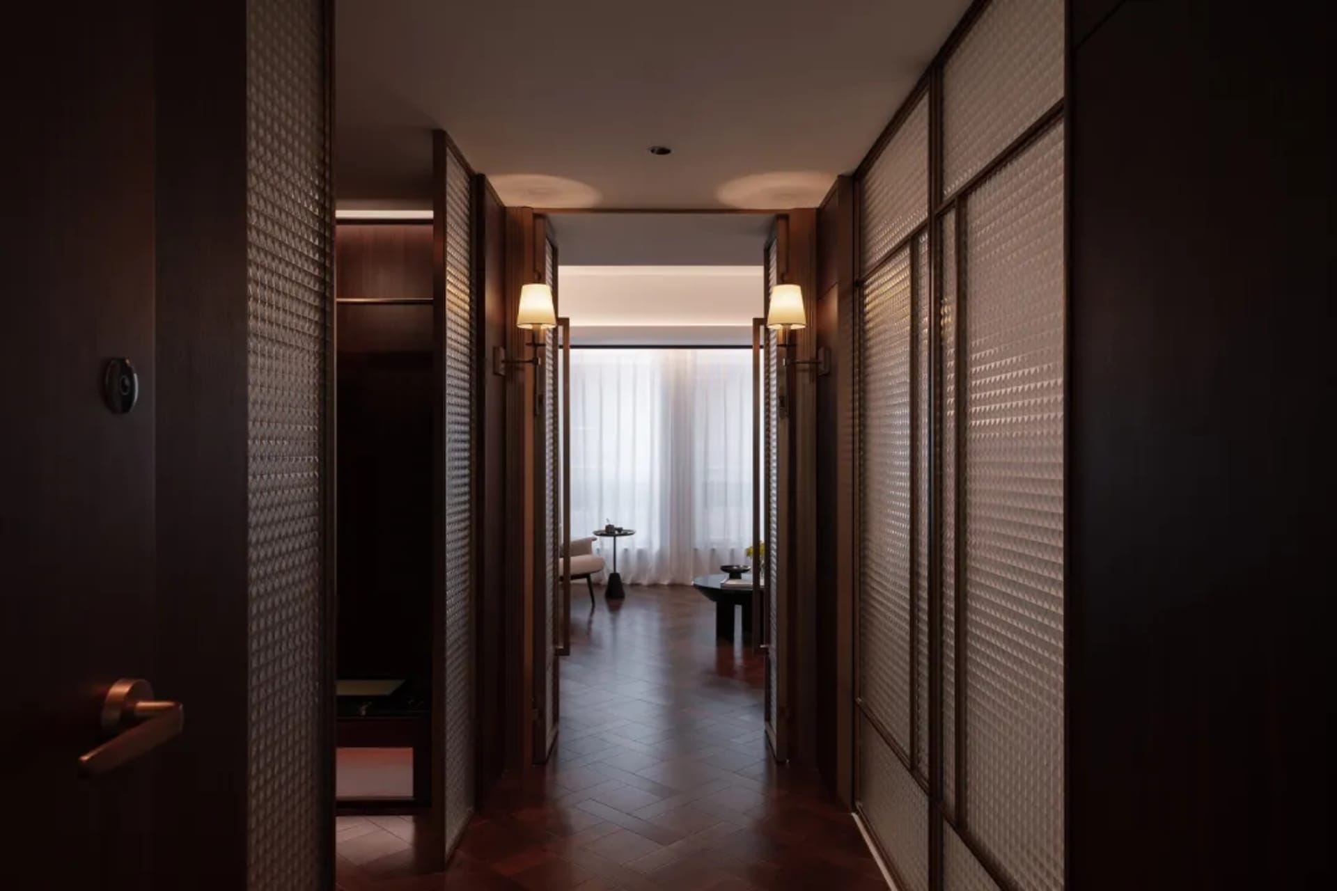 Dimly lit hotel hallway with a wooden floor, a glass door, a couch, a chair, and a vase on a surface
