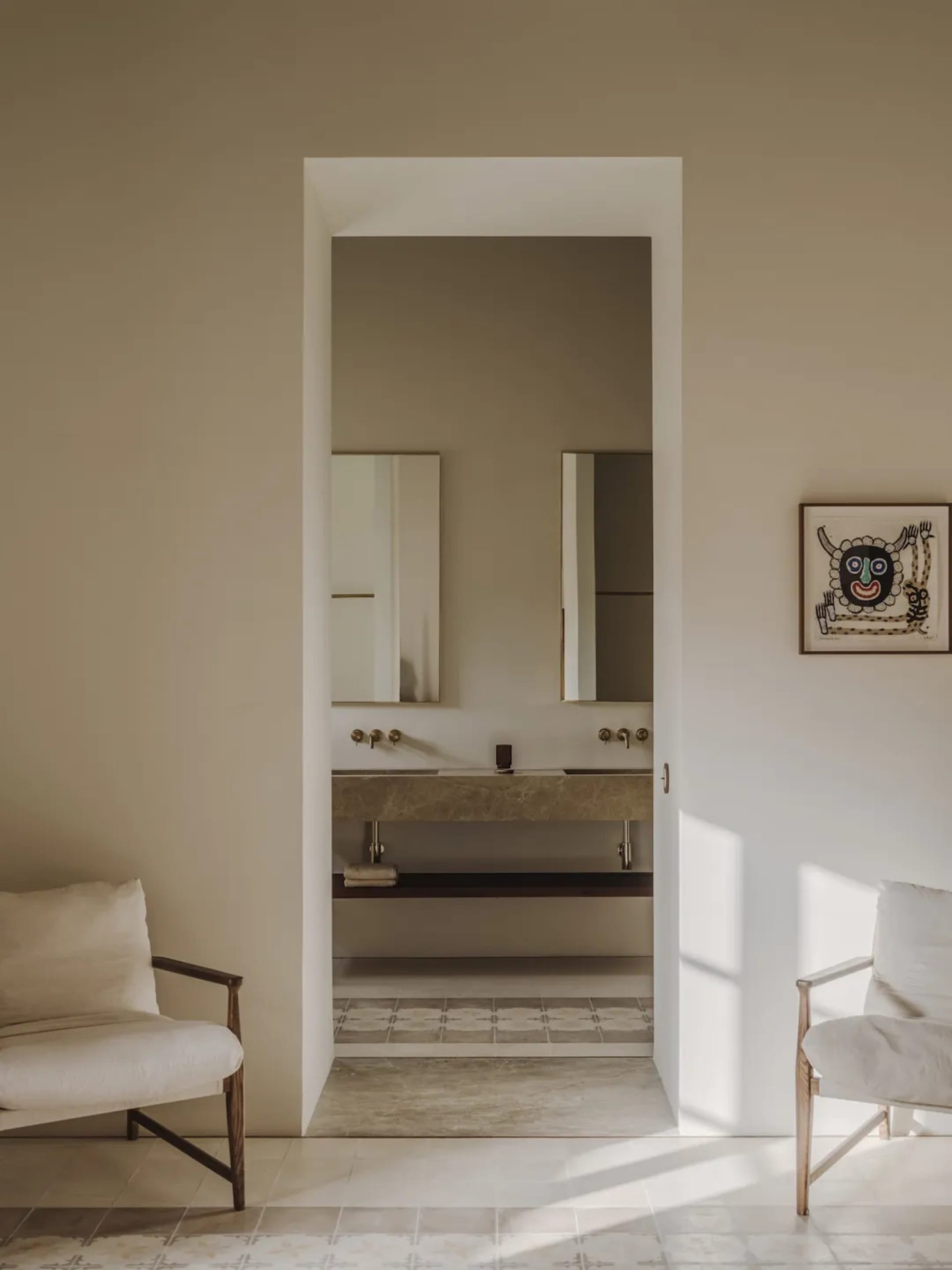A clean and organized residential bathroom with a white tile floor, sink, mirror, and chair, lit by natural sunlight