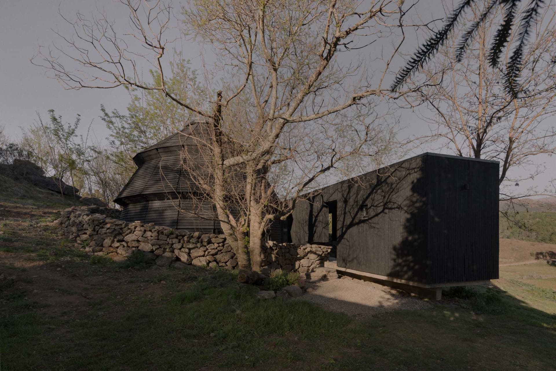 A black house with a stone wall surrounded by trees, situated on a hill, creating a nostalgic atmosphere