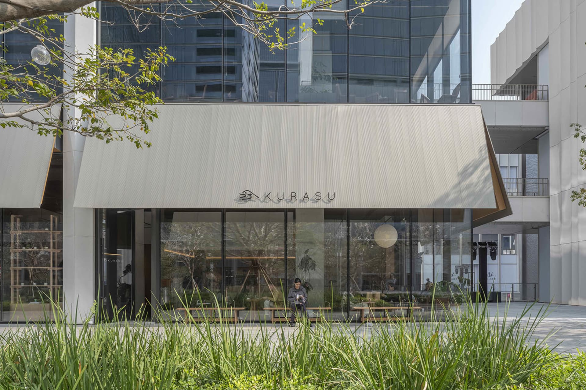A restaurant building with a large window, a sign, and a tree outside, set in a neutral color palette with natural light pouring in