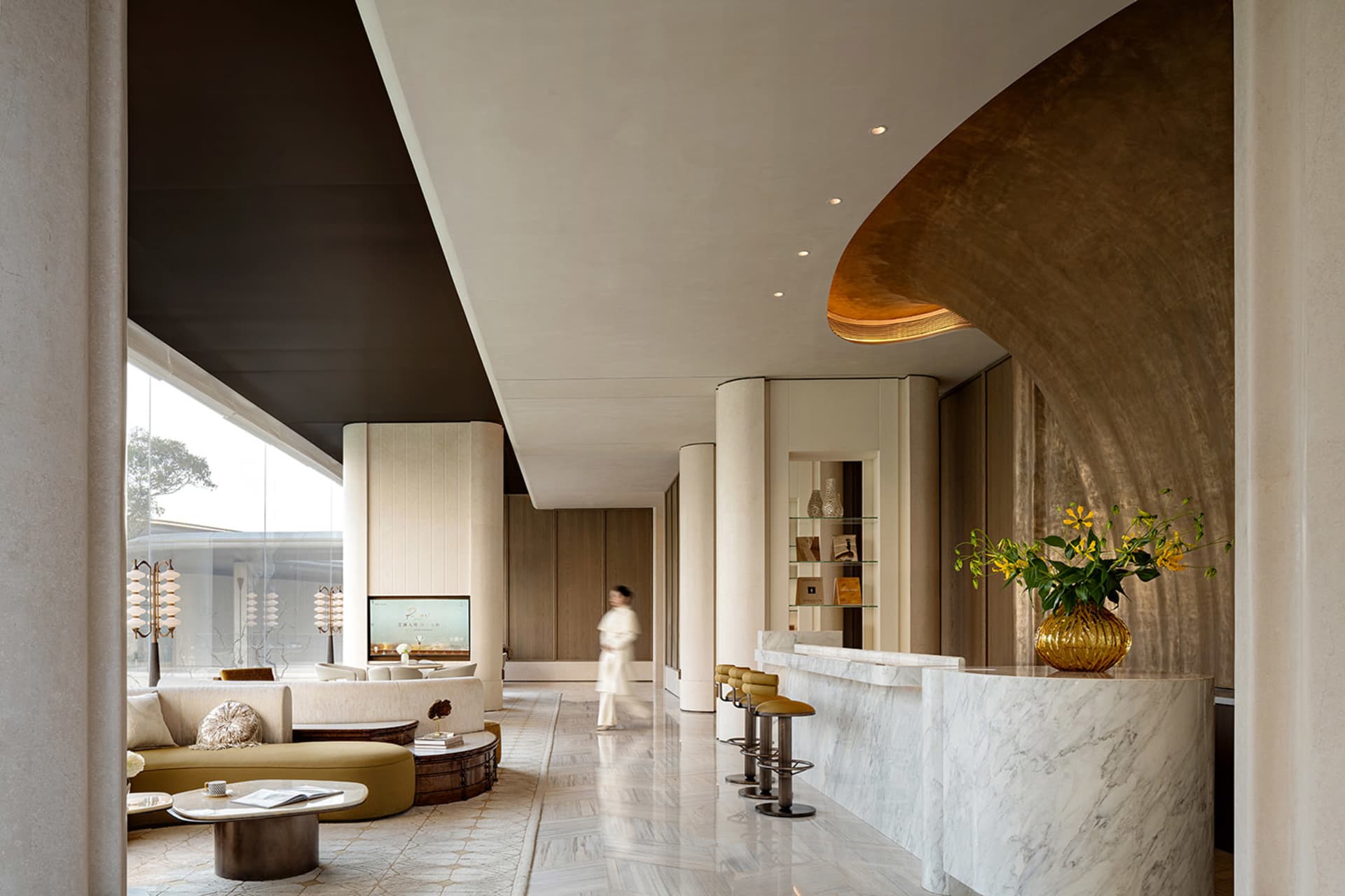 Well-lit hotel lobby with a modern design, featuring a large counter, couch, and dining table, with a woman walking through, and natural light streaming in through the windows