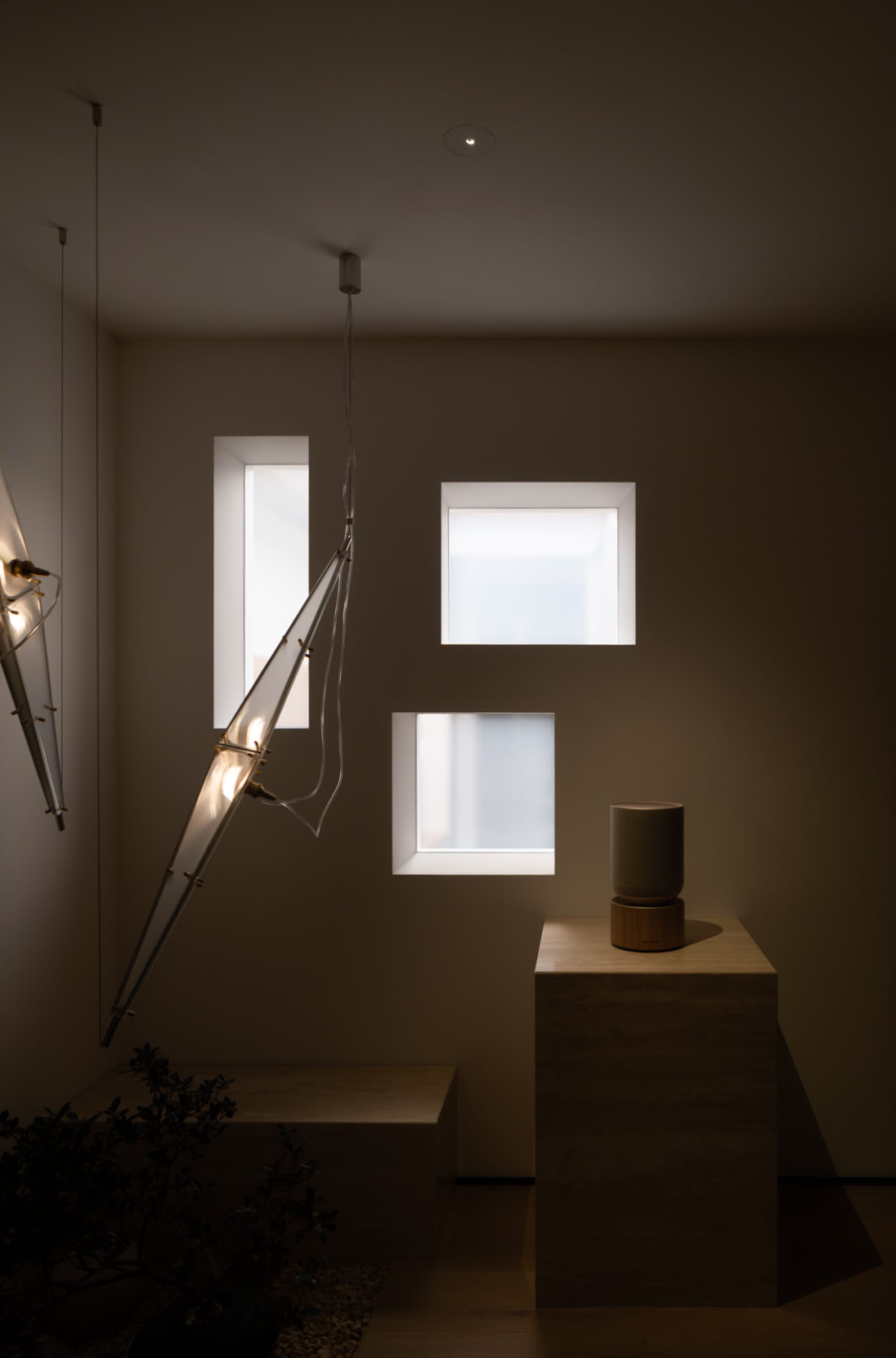 Small dimly lit room with a white wall, a window, two hanging lights, a table with a vase, and a potted plant near the left side