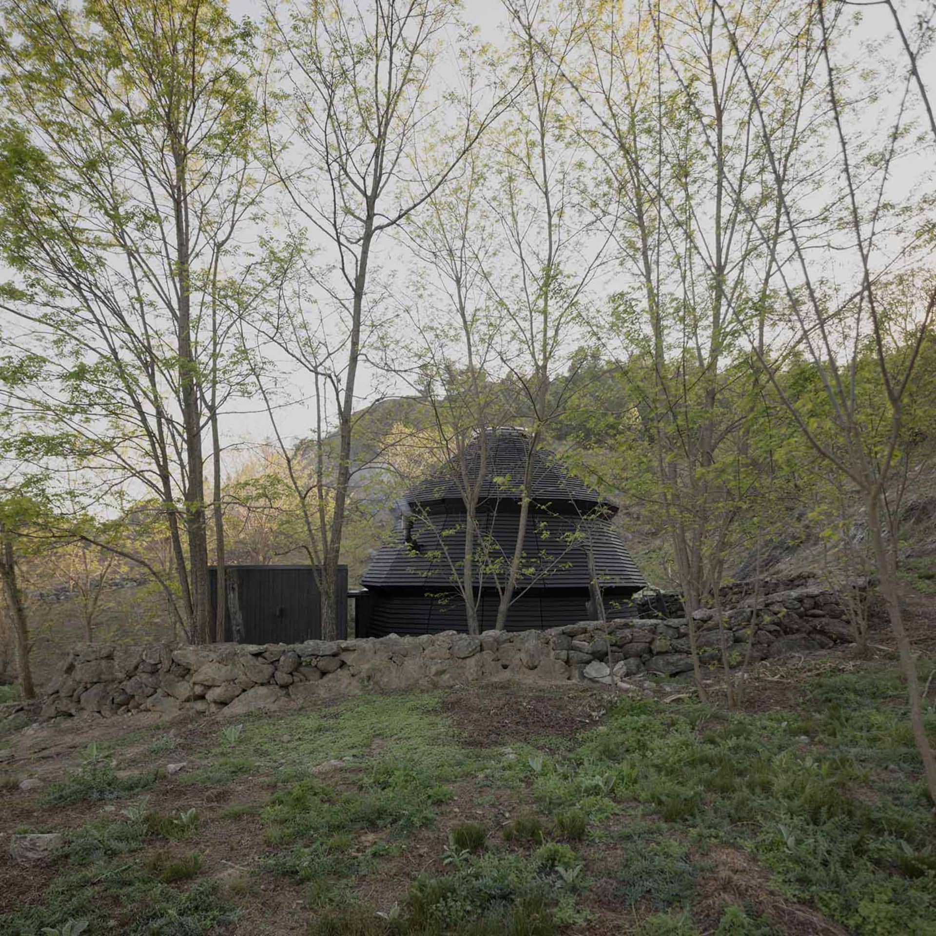A small house or shed with a stone wall and black roof, set in a grassy area surrounded by trees