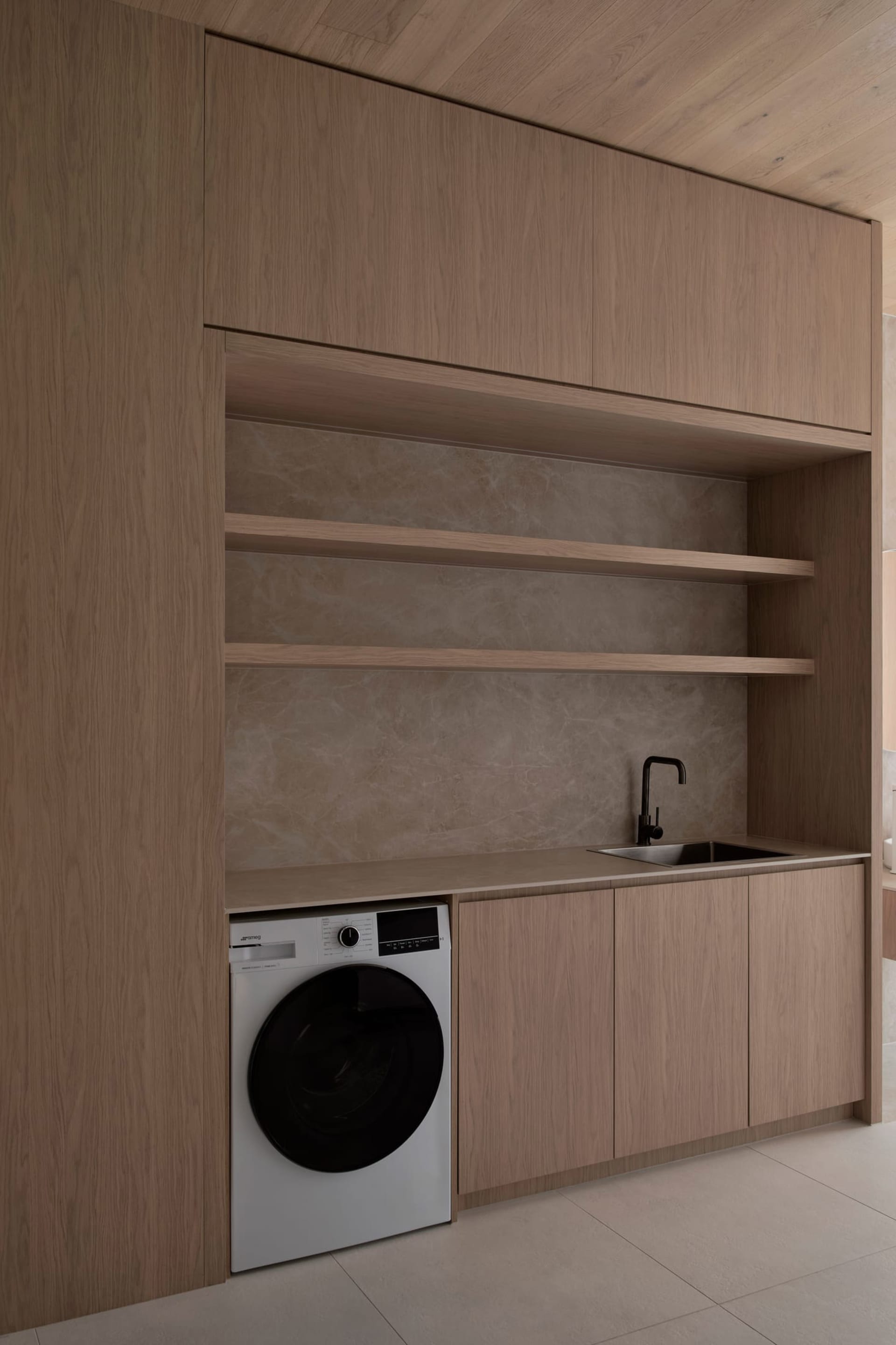 A modern kitchen with wooden cabinets, a white washer dryer, and a refrigerator, sink, and microwave, lit by natural sunlight