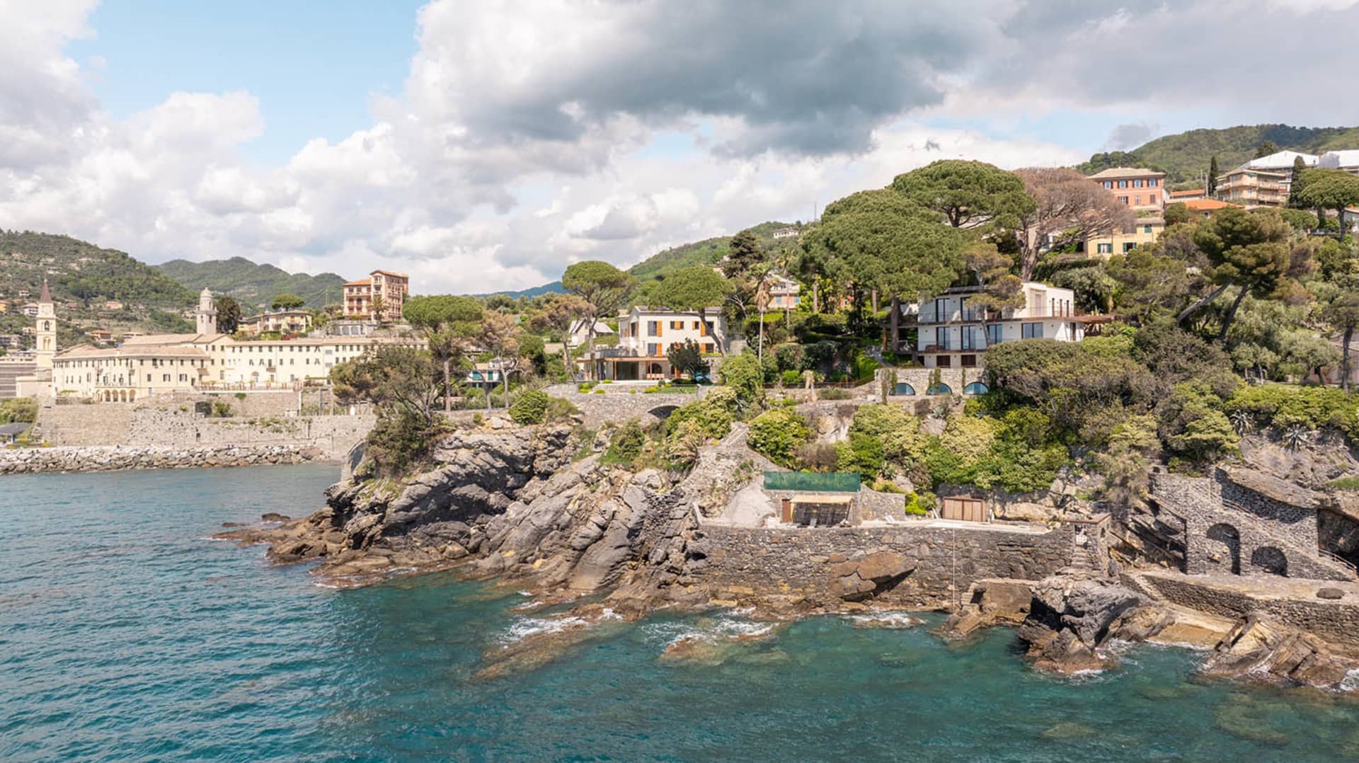 A residential area with a mix of modern and traditional houses on a hillside, surrounded by lush greenery and overlooking the water